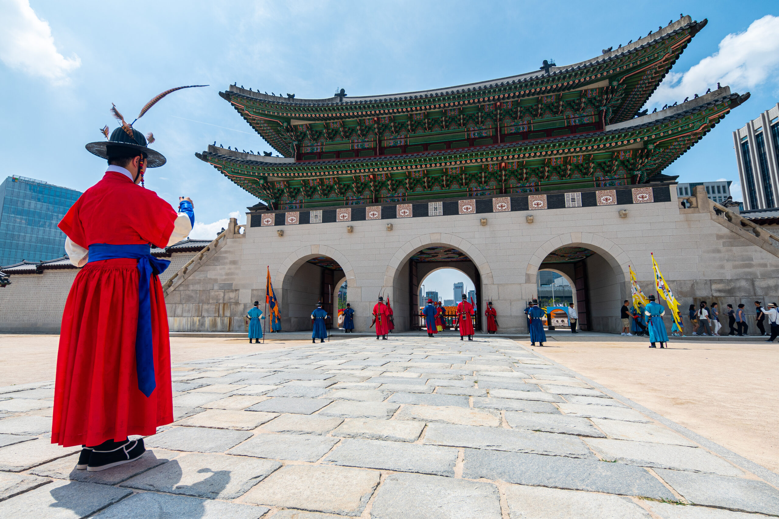 Person in a bright red traditional hanbok stands at a historic ornate gate with a colorful eaved roof on a sunny day; several people in period clothing are nearby.