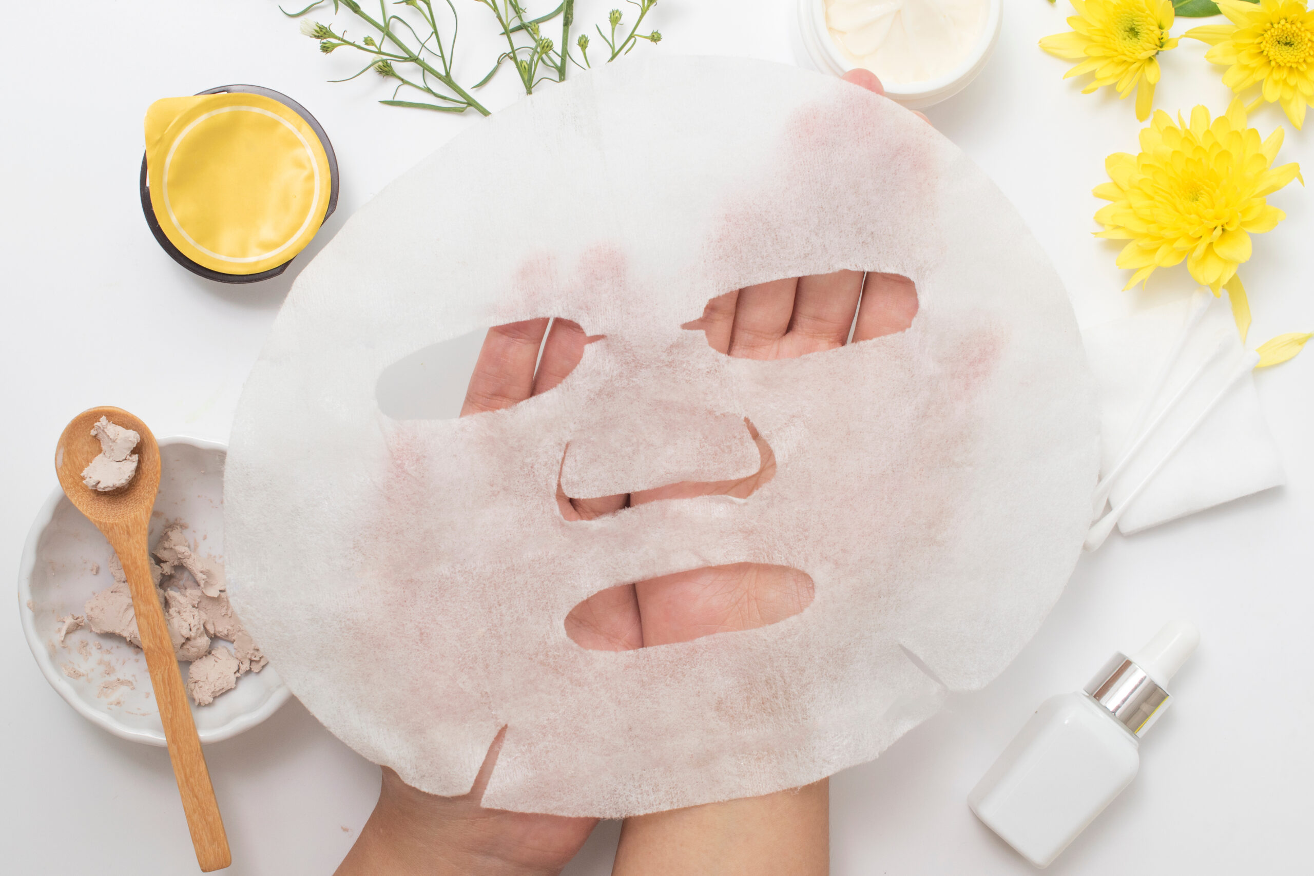 Hands holding a torn sheet facial mask over a spa-inspired setup with yellow flowers and skincare items nearby, viewed top-down.