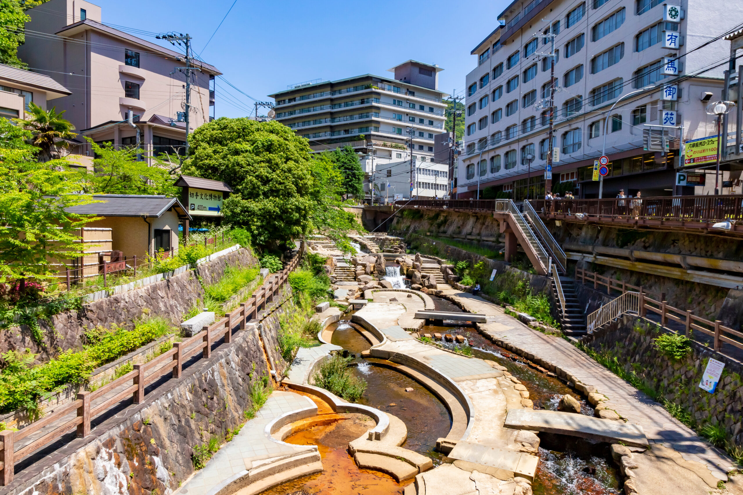 Urban riverscape with stepped concrete channels, wooden railing, and green trees beside sidewalked pedestrian path near multi-story buildings on a sunny day in Japan.