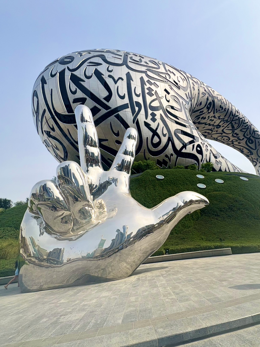 Large polished metal hand making a peace sign, in front of a curved sculpture covered in Arabic calligraphy.