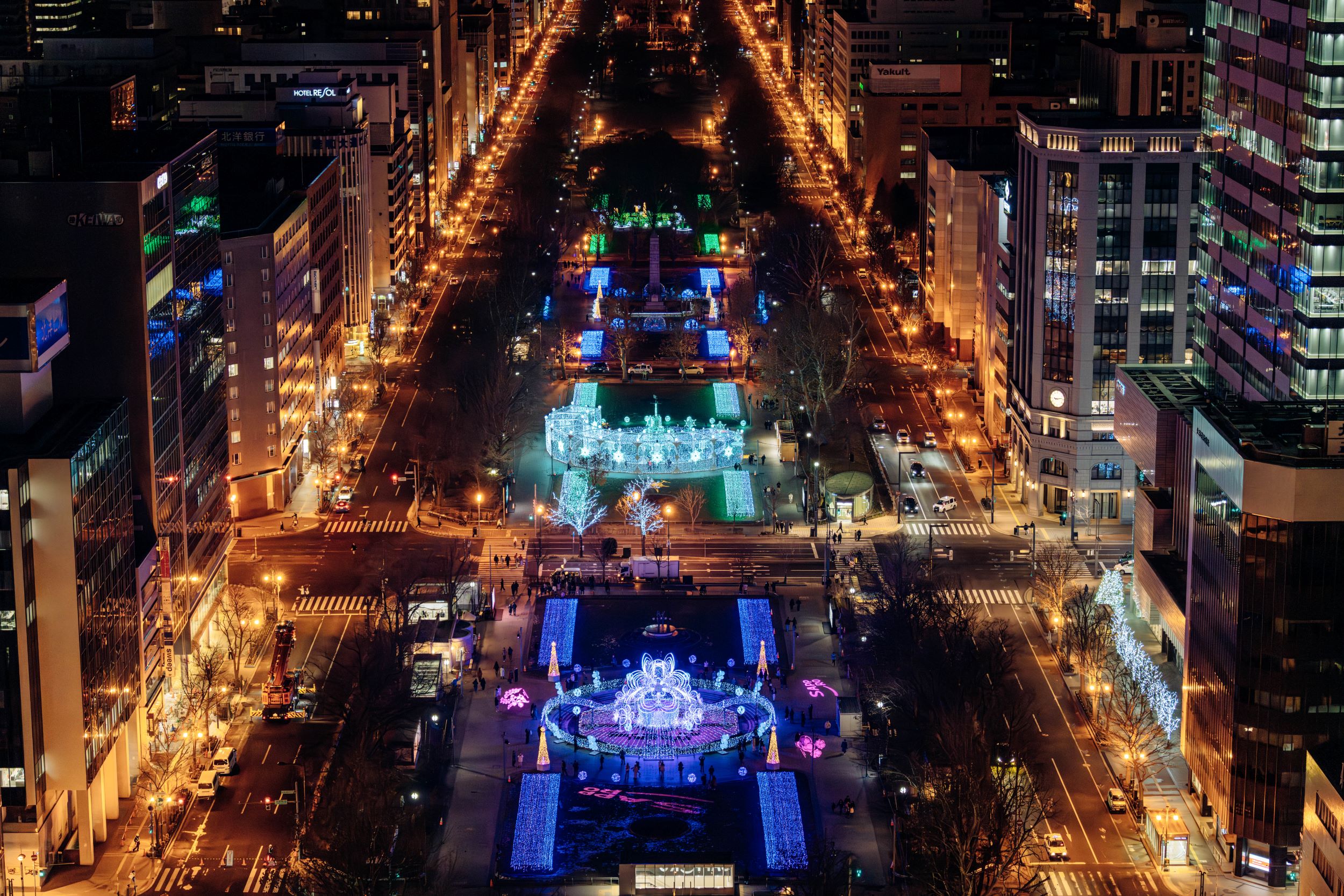 Aerial nighttime view of a city center featuring a central park with blue decorative lights, a fountain area, and surrounding illuminated streets and tall buildings.