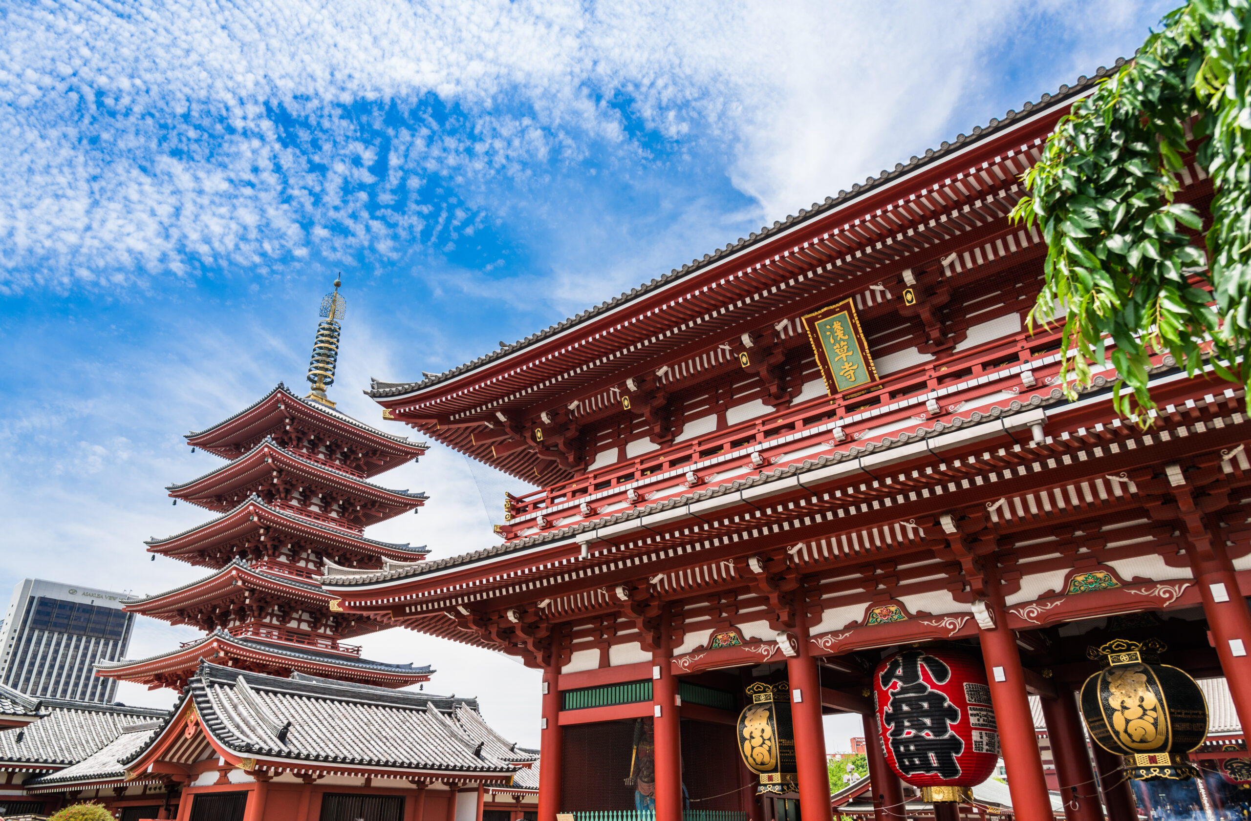 Red wooden temple complex with tiered pagoda, hanging lanterns, and ornate rooflines against a bright blue sky with green foliage nearby.