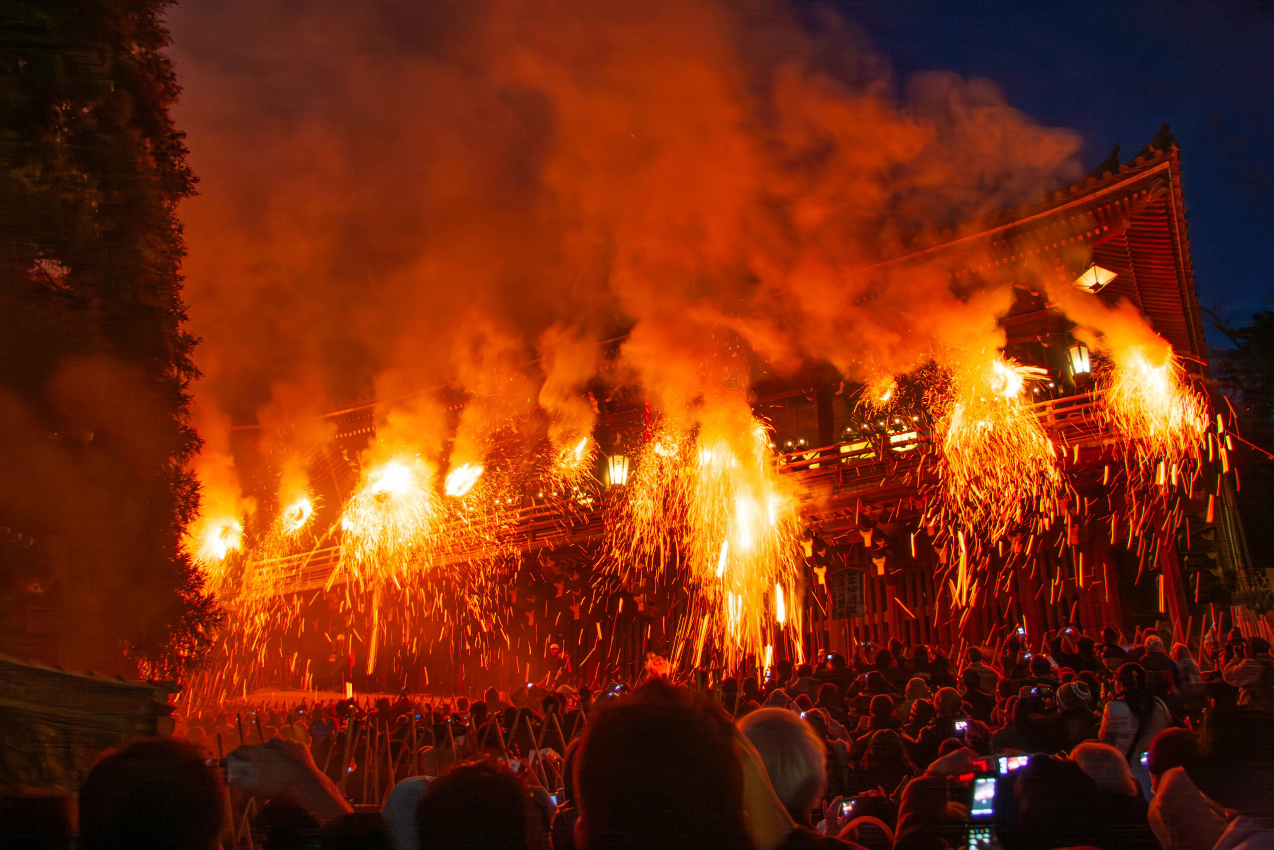 Night festival scene with a crowd watching as large orange flames and sparks erupt from torches along a wooden, traditional building at dusk/dawn lighting the area.