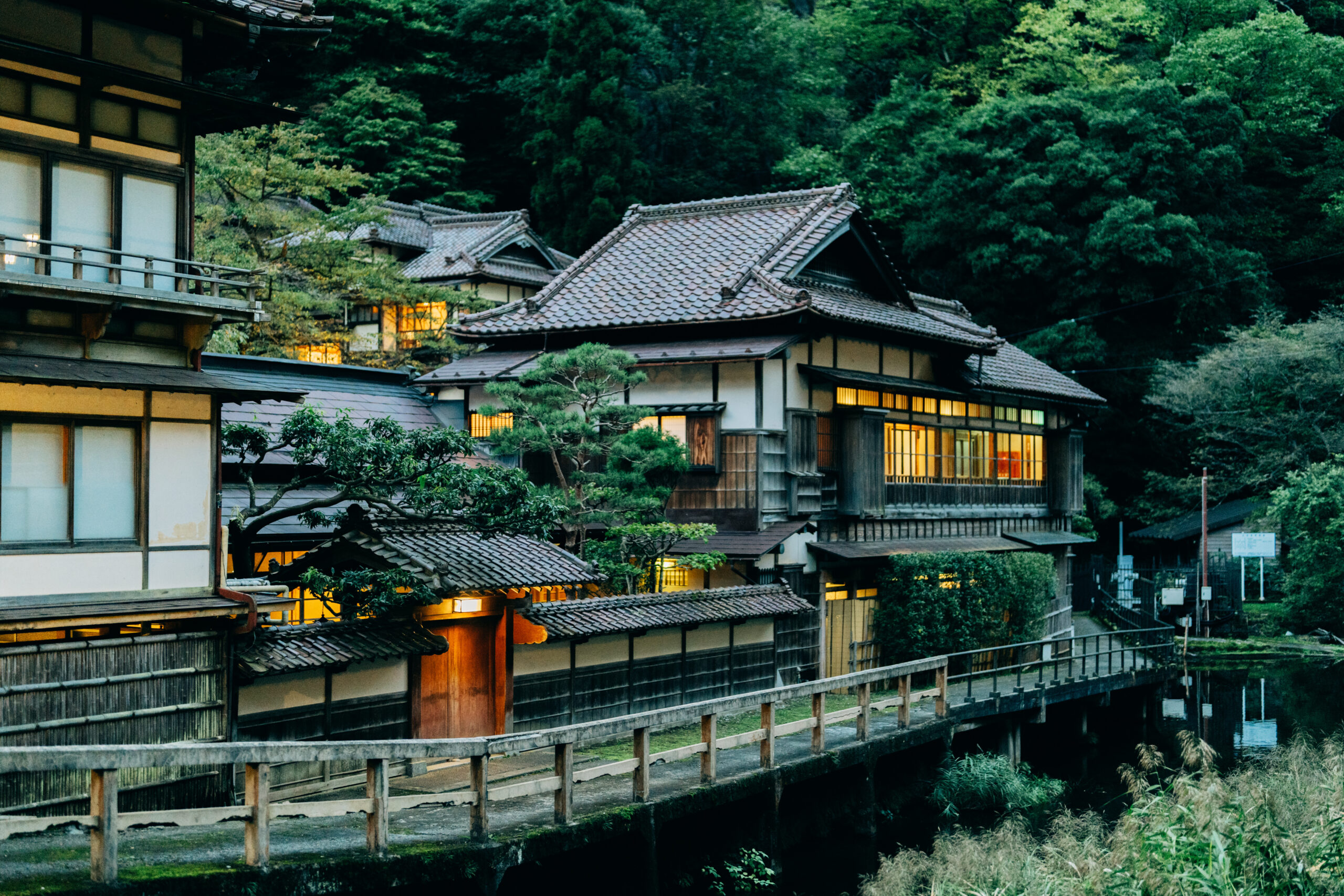 Traditional Japanese wooden houses with tiled roofs along a calm canal, framed by green hills and trees at dusk.