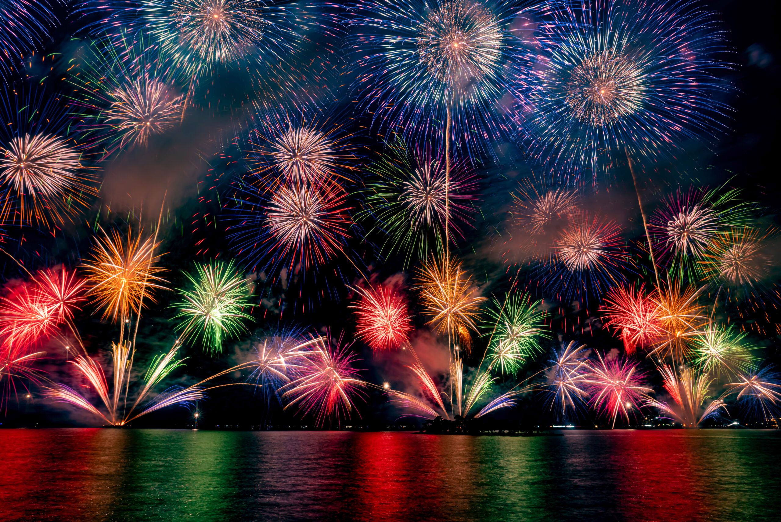 Night scene of colorful fireworks exploding over a calm body of water with vivid reflections.