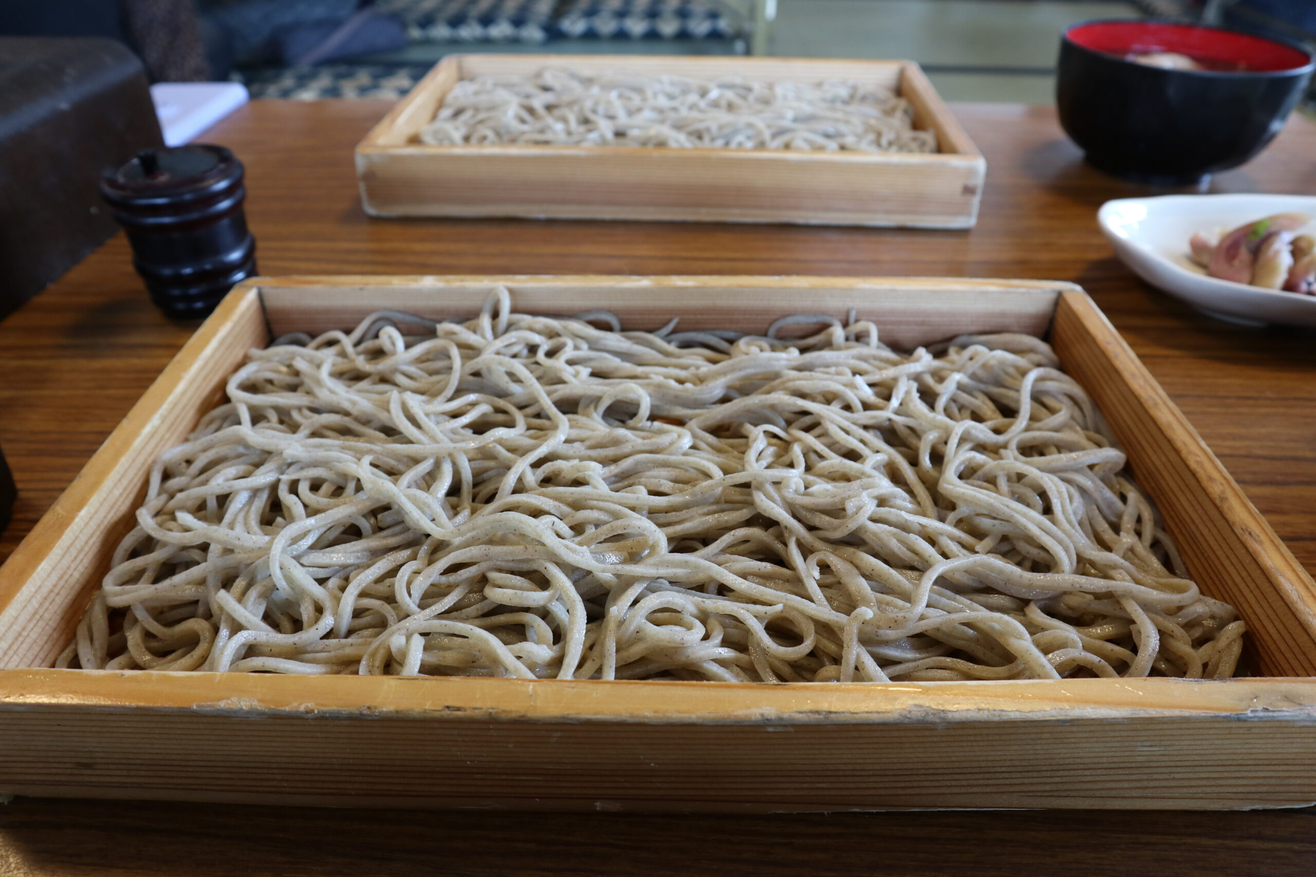 Wooden tray filled with gray soba noodles on a wooden table, with a dark sauce cup nearby.