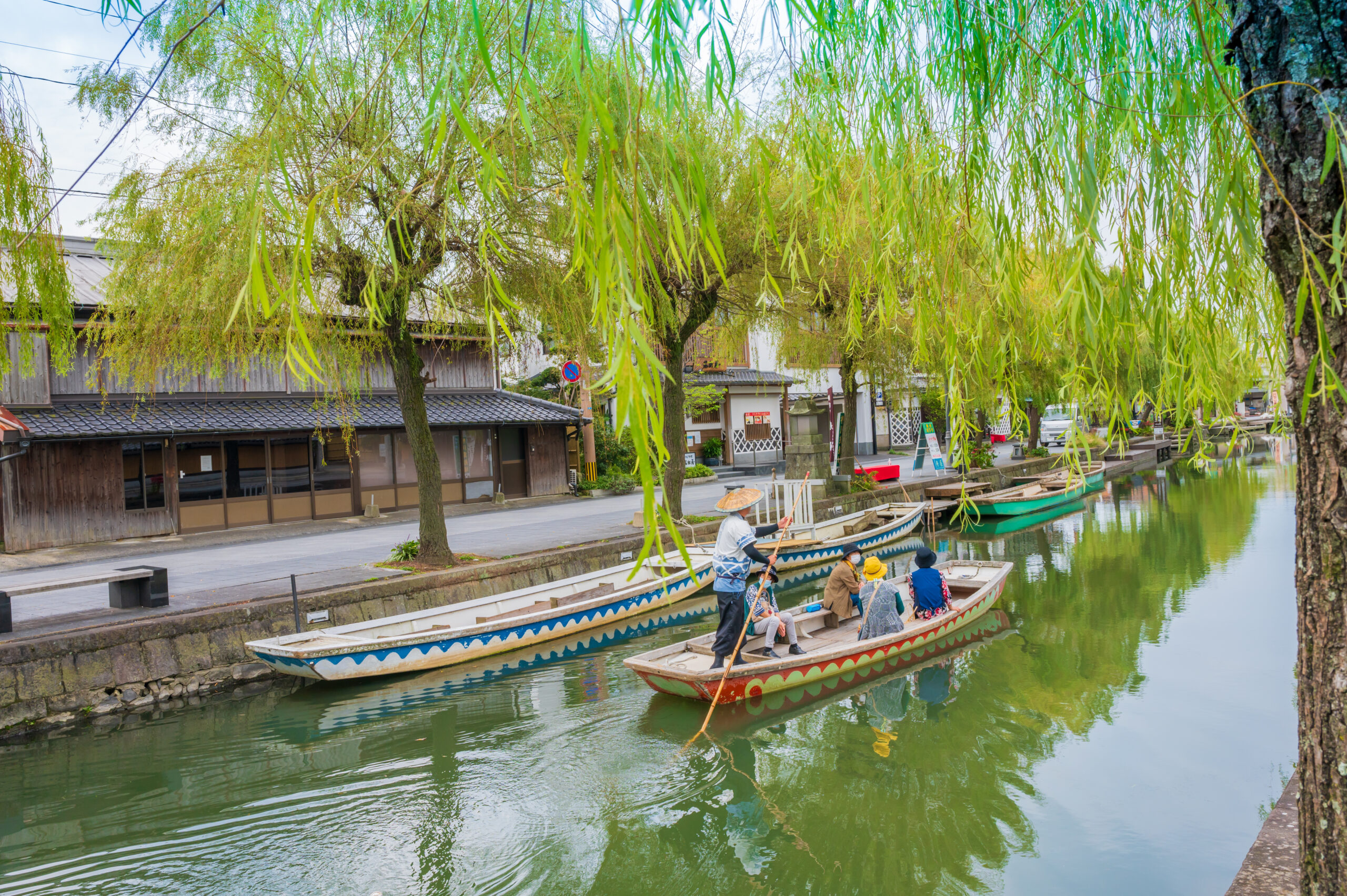 Group of people paddling a long decorative boat along a canalside, with hanging willow branches overhead and traditional wooden buildings nearby.