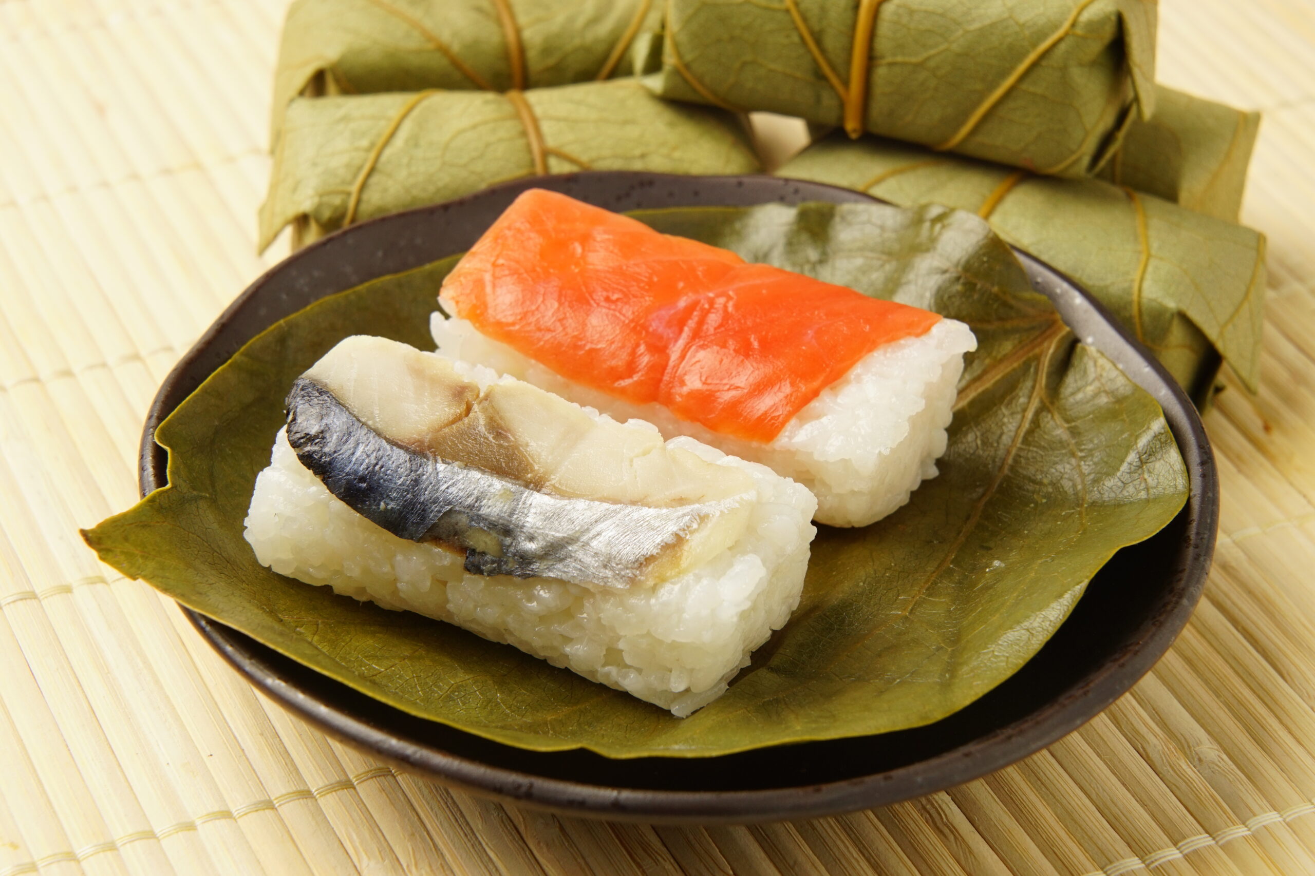 Two nigiri sushi pieces (salmon and mackerel) on a green leaf, served on a dark plate with a bamboo mat background.