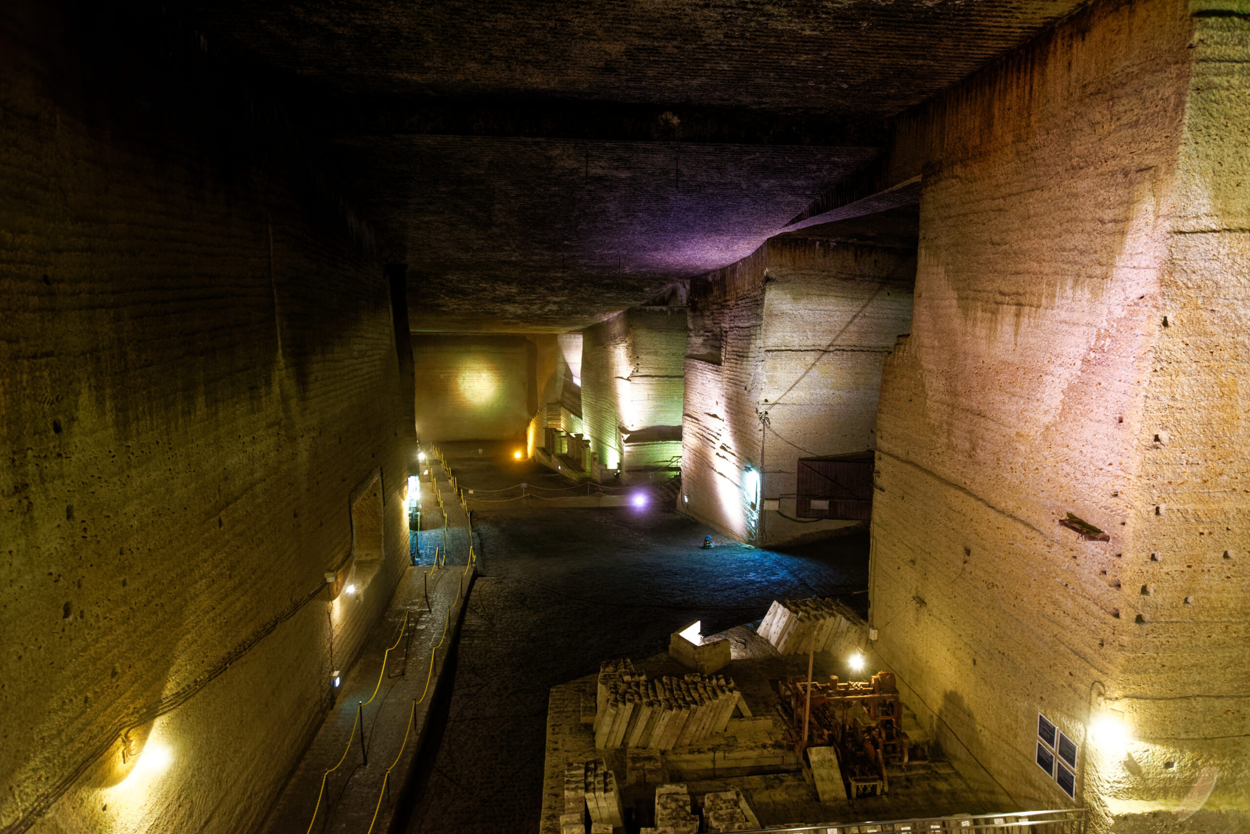Inside a vast underground quarry cavern with tall rocky walls, lit by colored lights and a walkway with railings at the center-right.