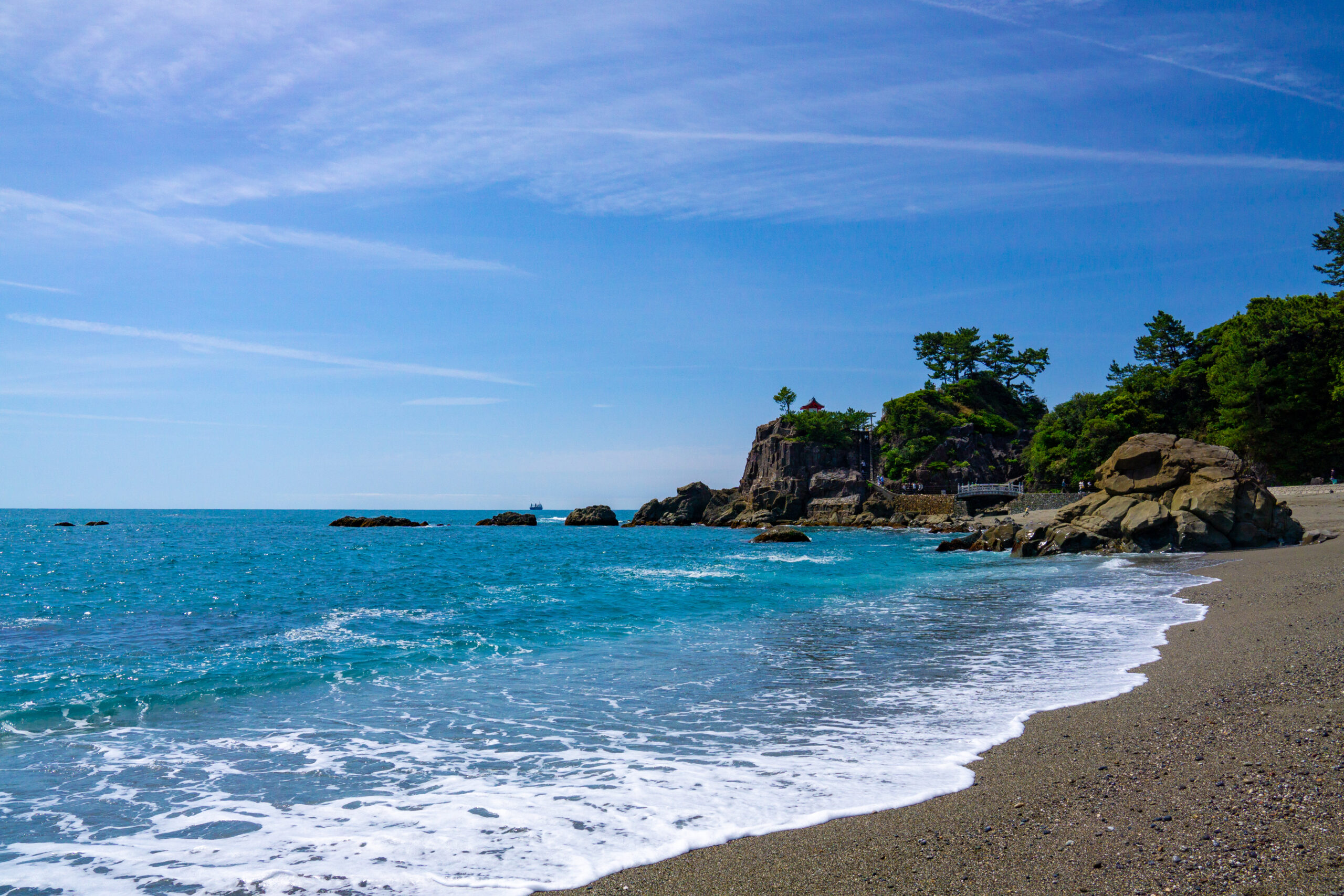 Coastal beach with blue sea, rocky headland on the right, and a small red pavilion among green trees at the top of the rocks