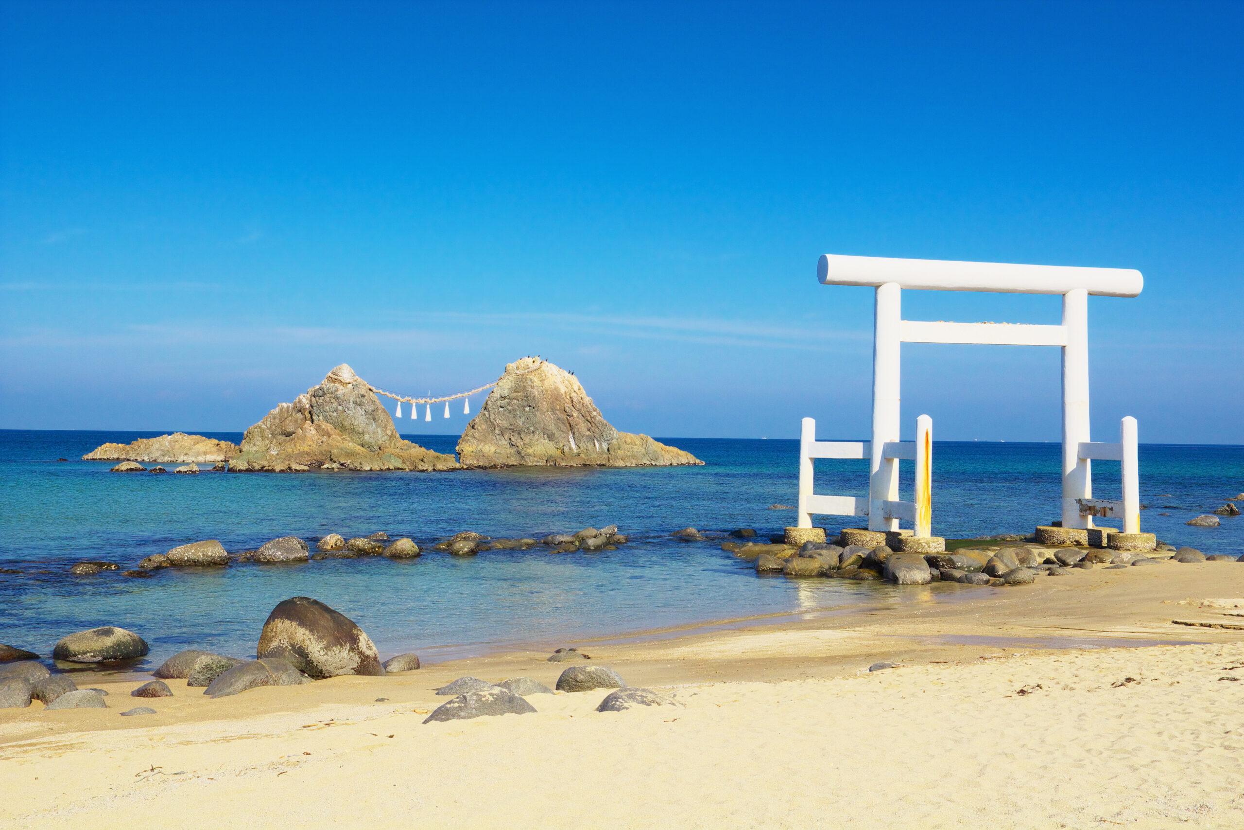 White torii gate by the coast with rocks in the foreground, a calm blue sea and clear sky behind it.
