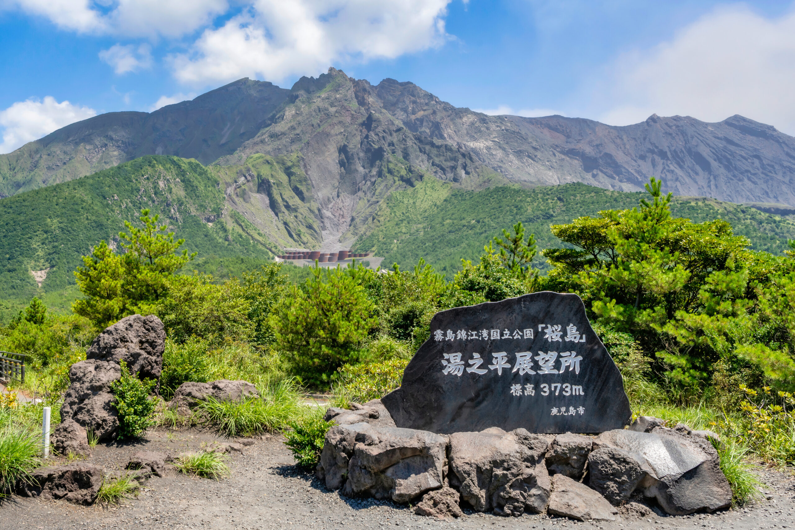 Mountain range with jagged rocky peaks and green slopes under a blue sky; in the foreground a dark stone plaque with white Japanese inscription, 373m.