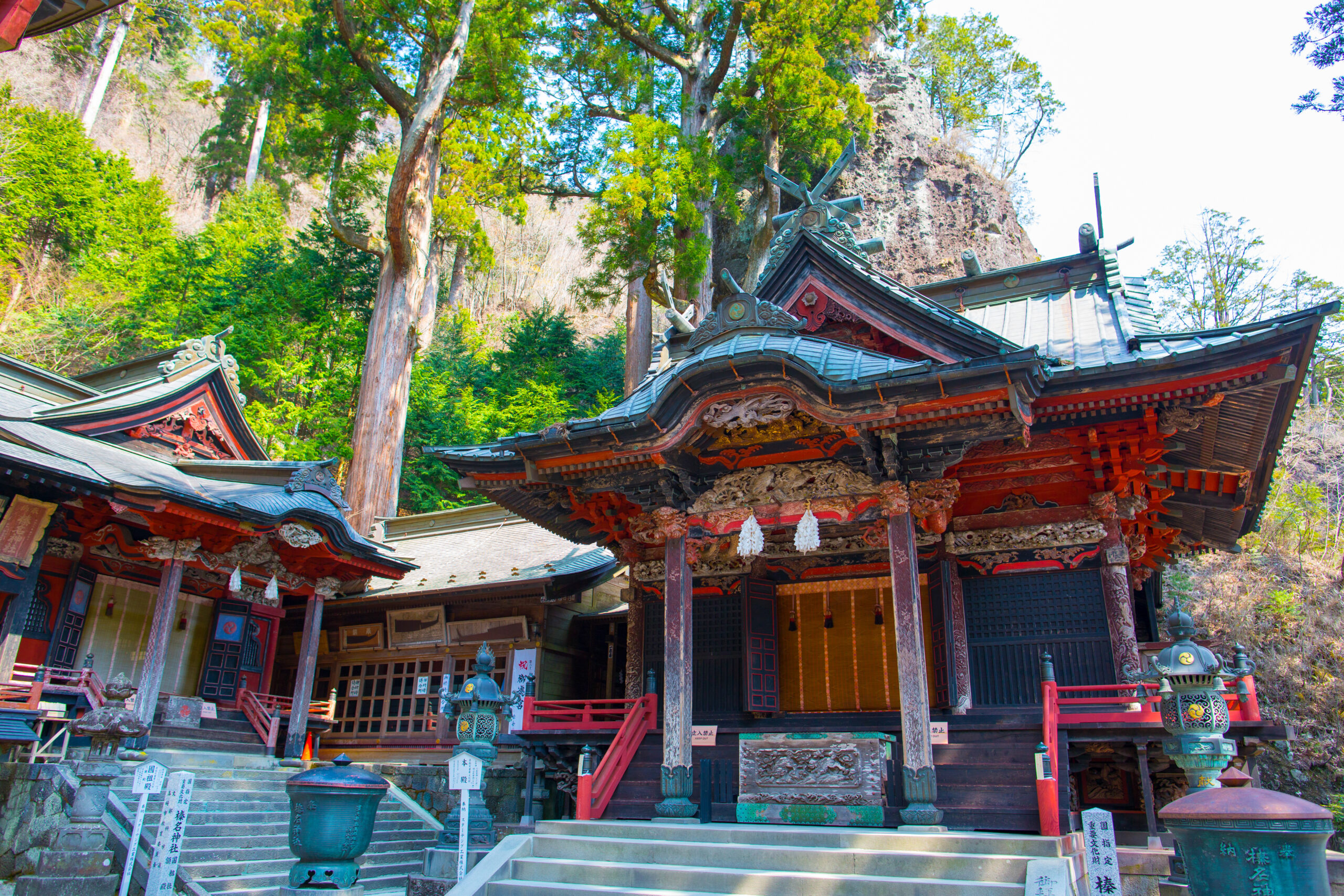 Colorful traditional temple with elaborately carved eaves set among green trees and a rocky cliff backdrop.