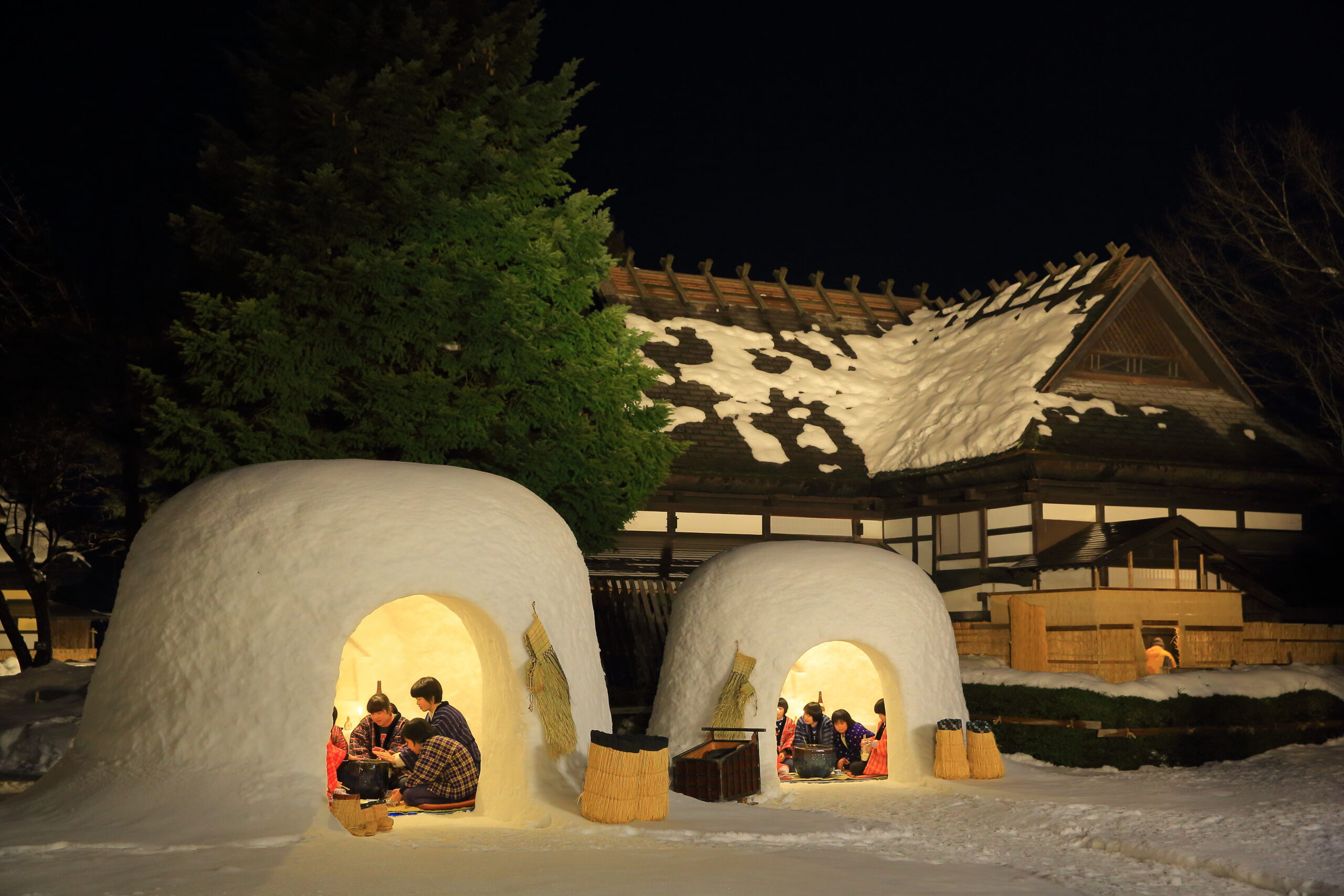 Nighttime scene of glowing snow igloos with people inside, set beside a wooden building and evergreen tree, under a dark sky.
