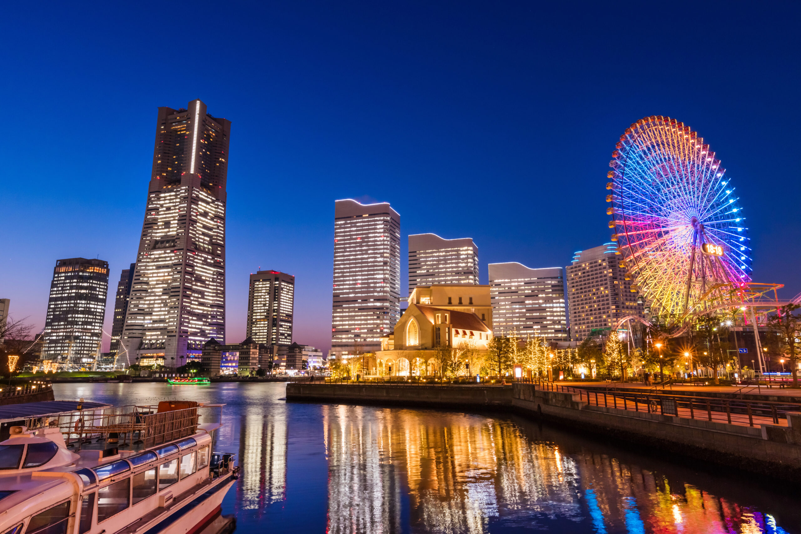 Night city skyline with tall illuminated buildings along a river and a colorful Ferris wheel on the right.