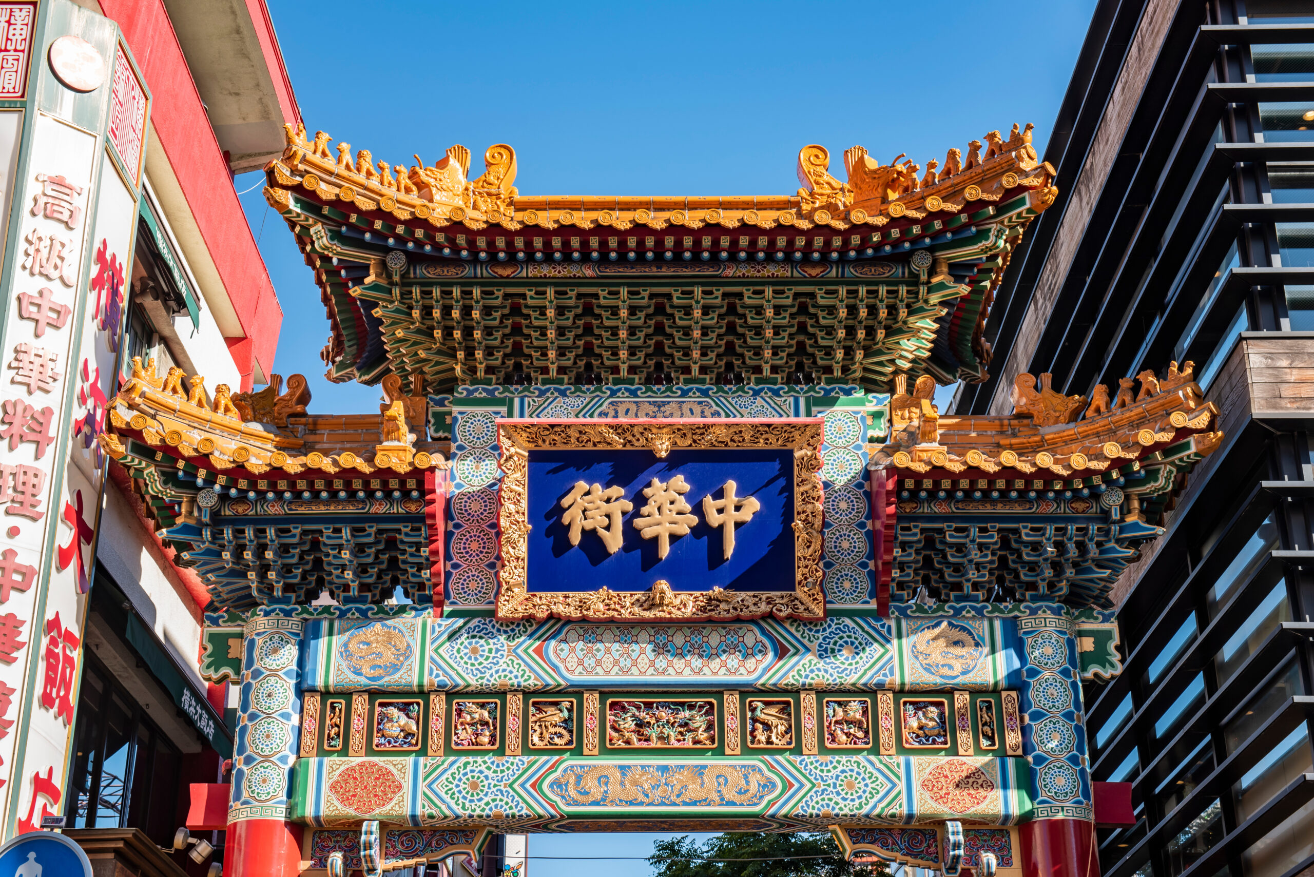 Colorful traditional Chinese archway with ornate gold roof decorations and a blue plaque bearing Chinese characters, between modern buildings on a sunny day edge-to-edge.