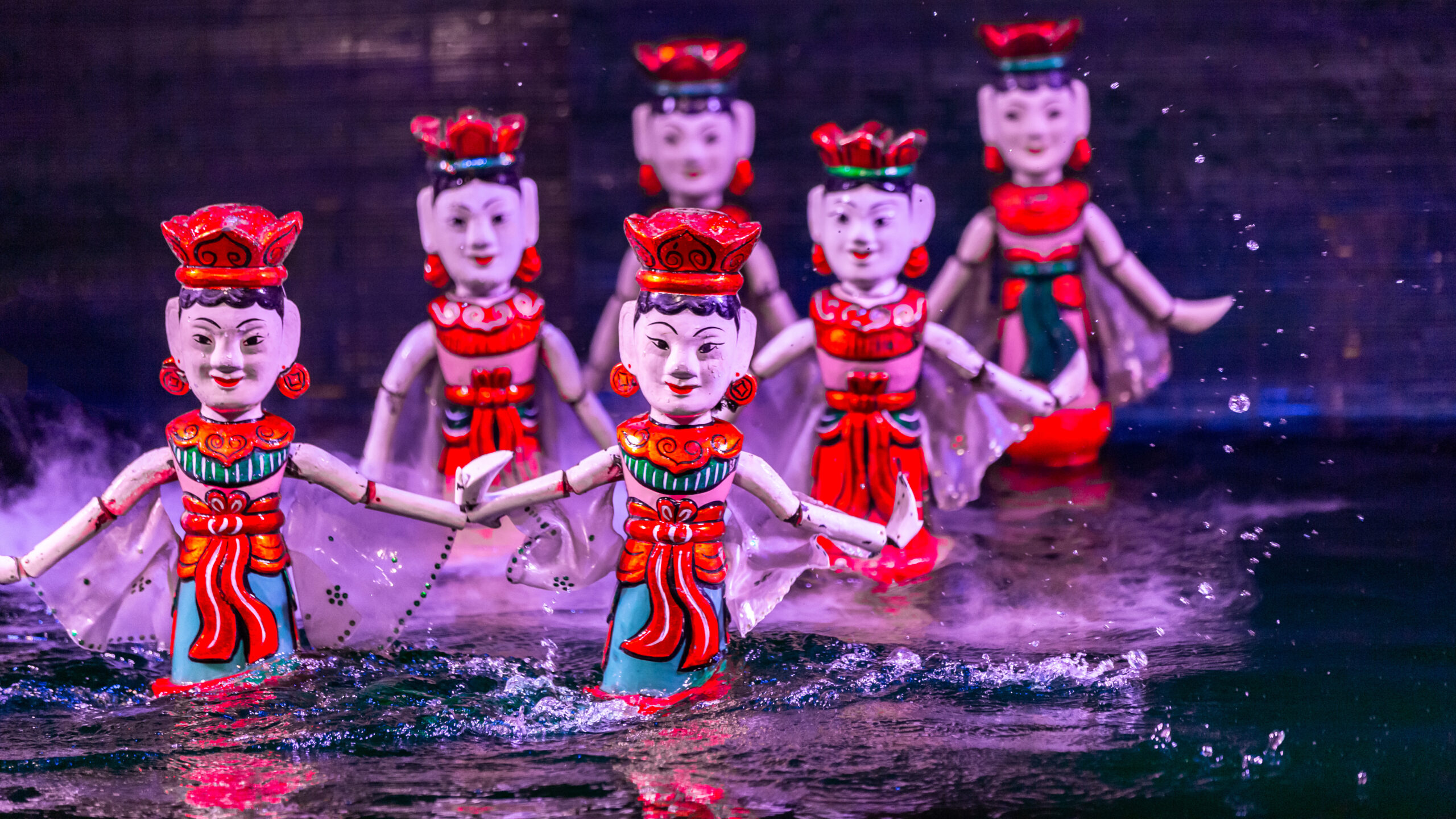 Colorfully painted water-stage puppets in red costumes performing a synchronized dance, with splashes around them.
