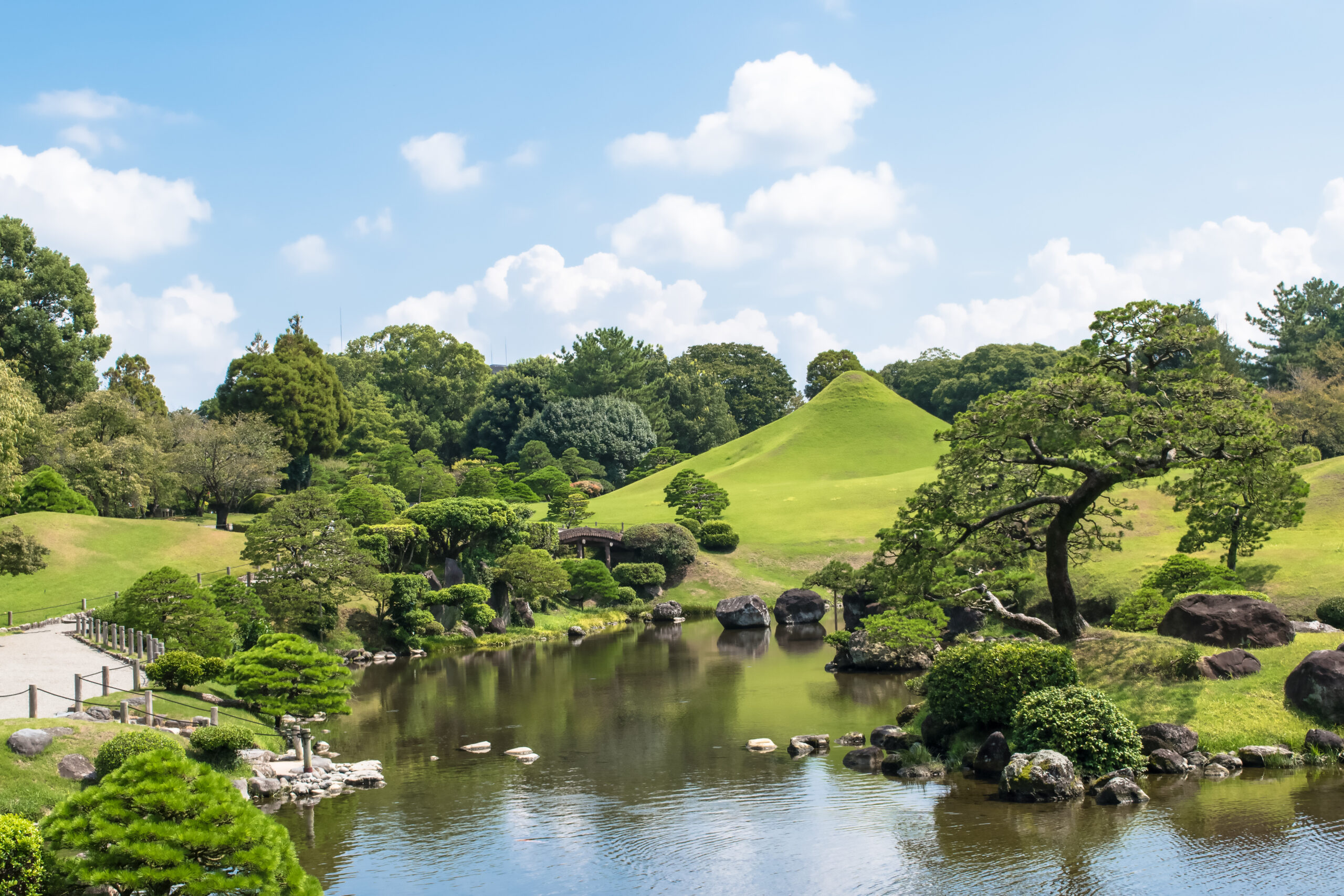 Panoramic view of a Japanese-style garden with a calm pond, trimmed trees, rocks along the shoreline, and a grassy hill under a blue sky.