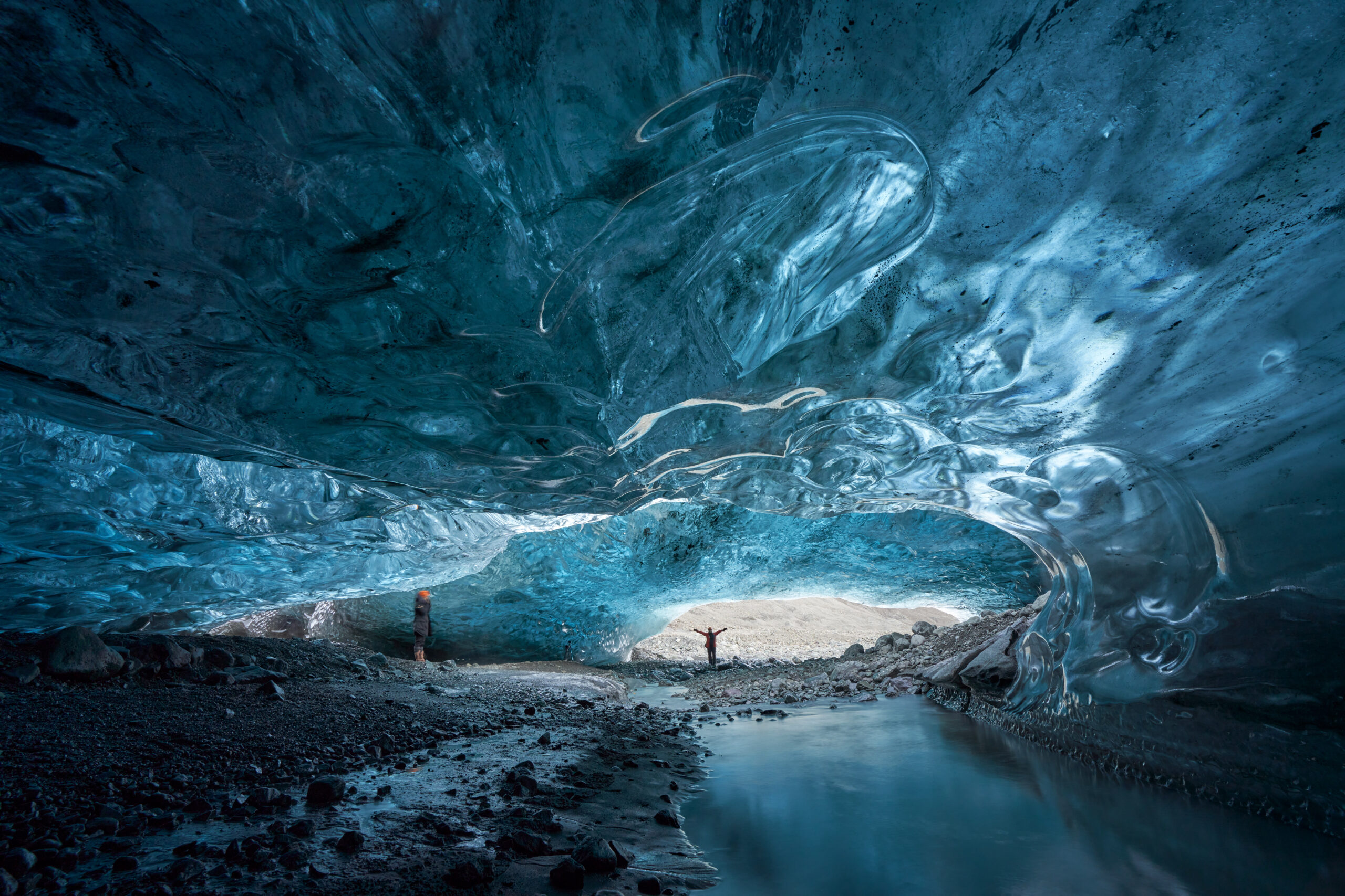 Inside a blue glacier ice cave, with sculpted walls and arches, two hikers standing on a rocky floor by a small pool of water.