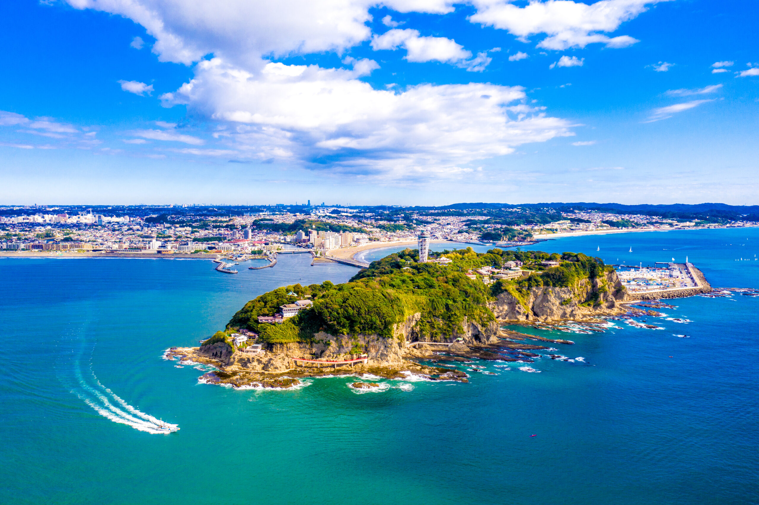 Aerial view of a coastal city with a harbor, beaches, and rocky promontories under a bright blue sky.