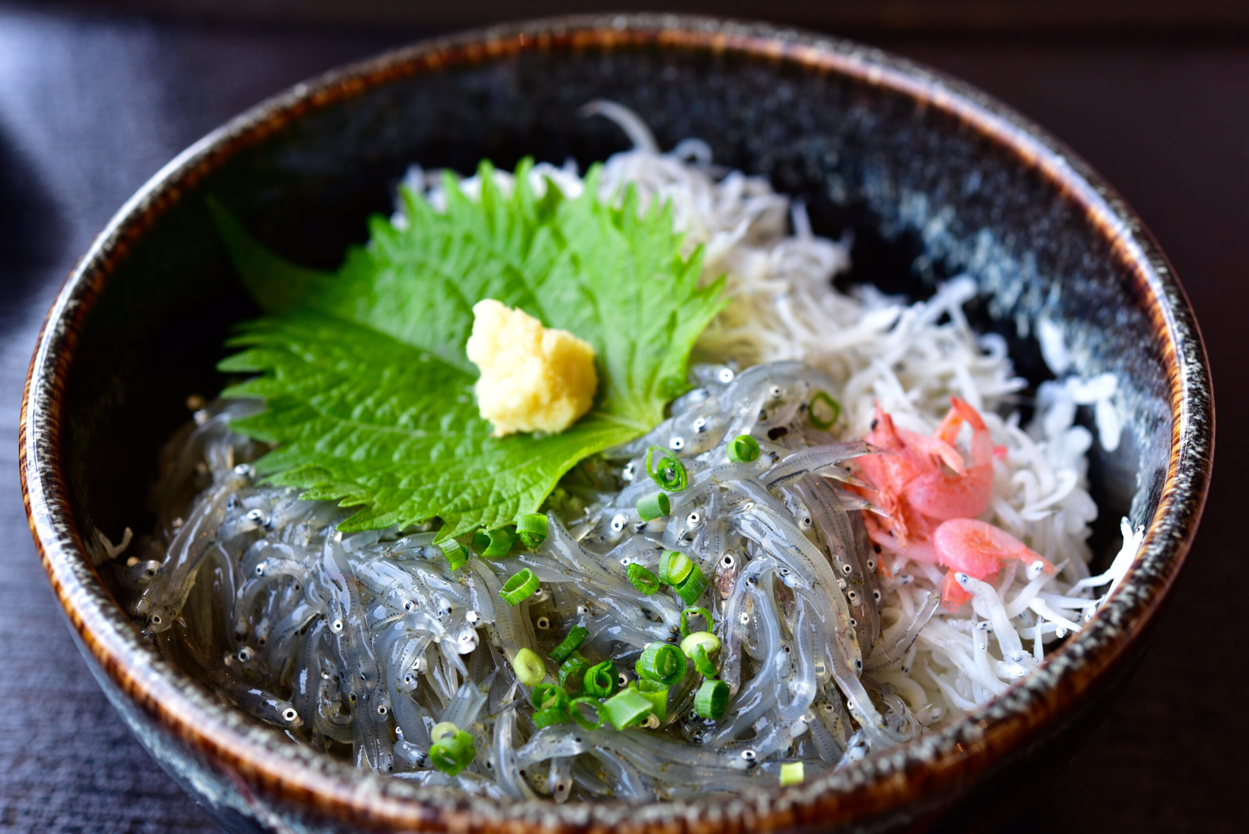 Bowl of tiny translucent shirasu fish over rice, topped with chopped green onions, a bright green shiso leaf, and grated ginger garnish.