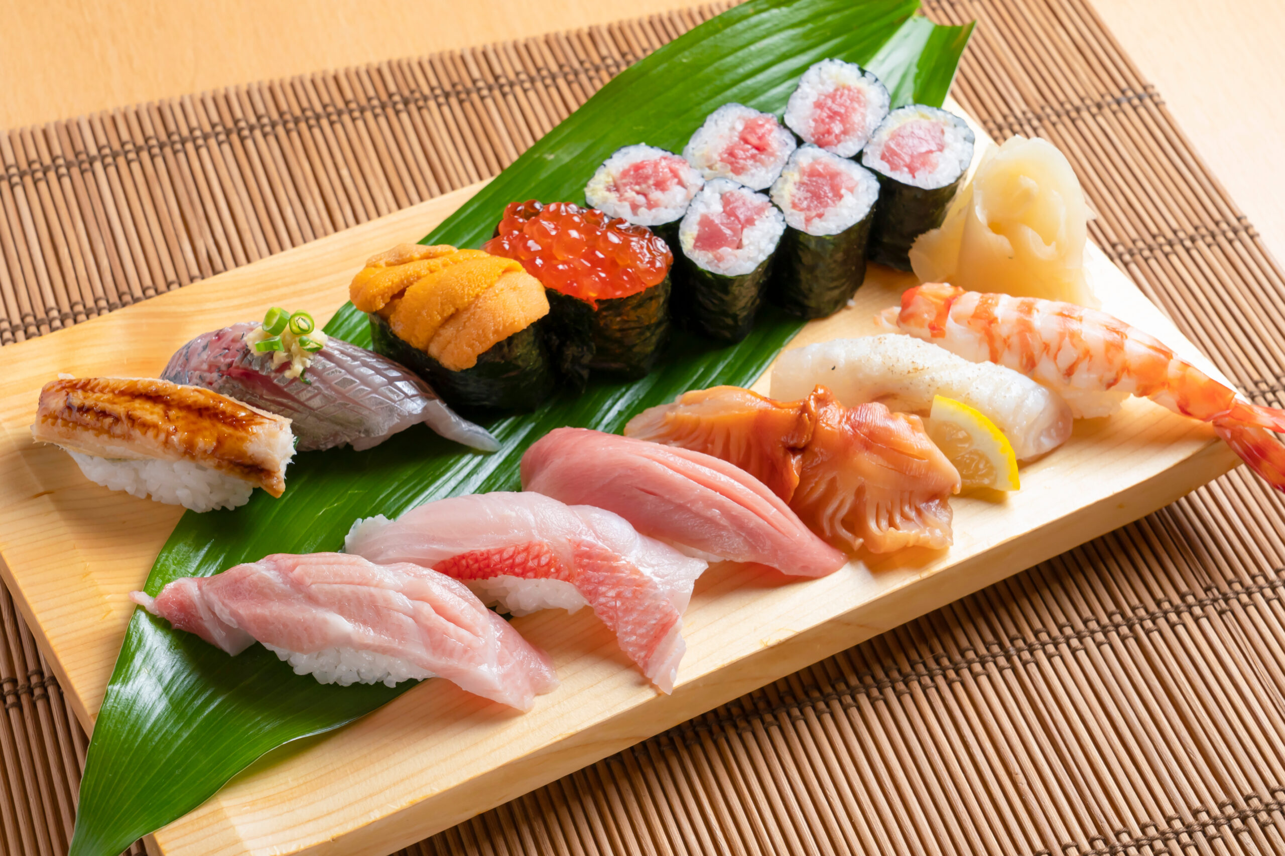 Assorted sushi on a green leaf, including nigiri and maki rolls, on a wooden tray.