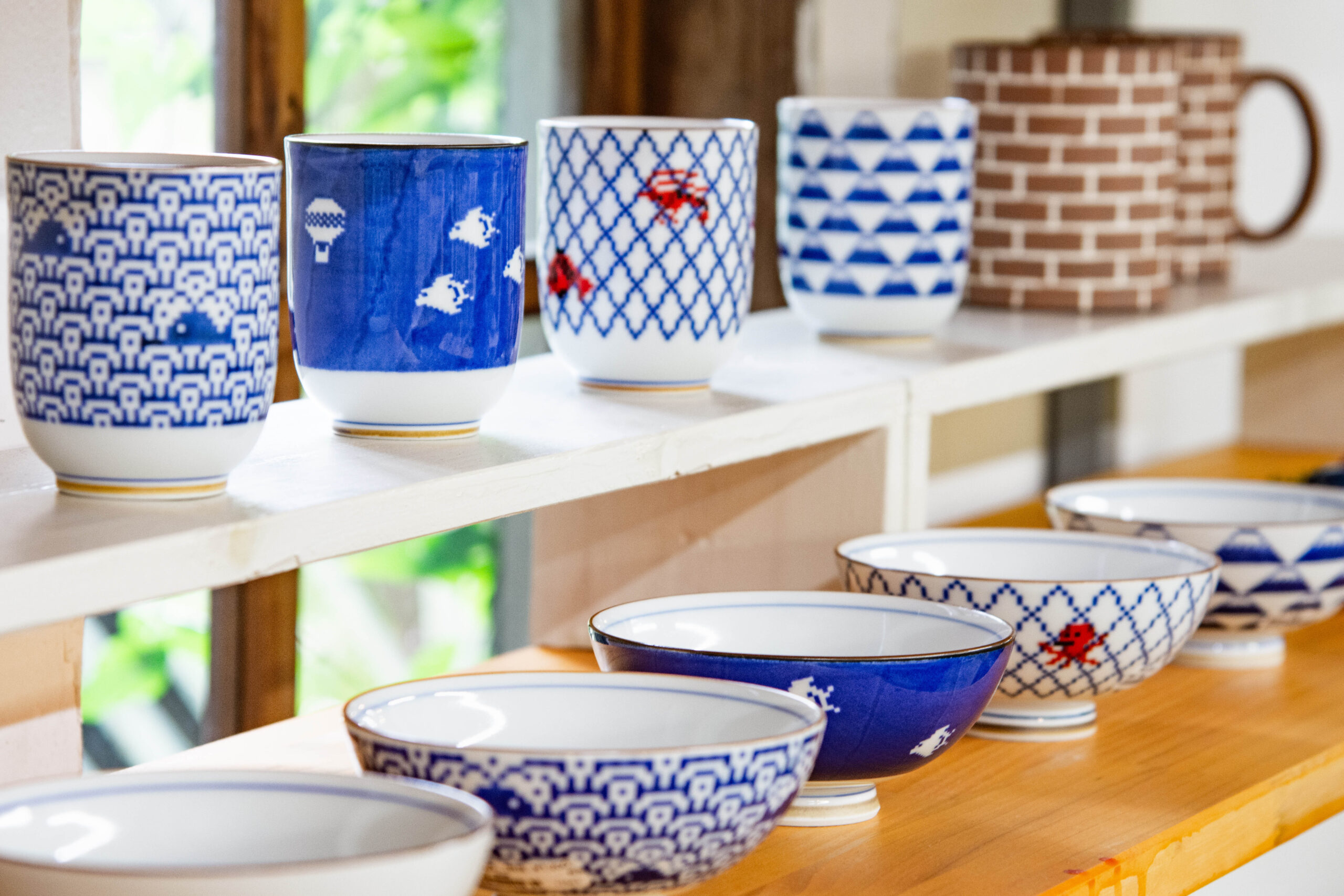 Row of blue and white patterned ceramic bowls and mugs lined up on a wooden shelf behind a kitchen/lurn area.