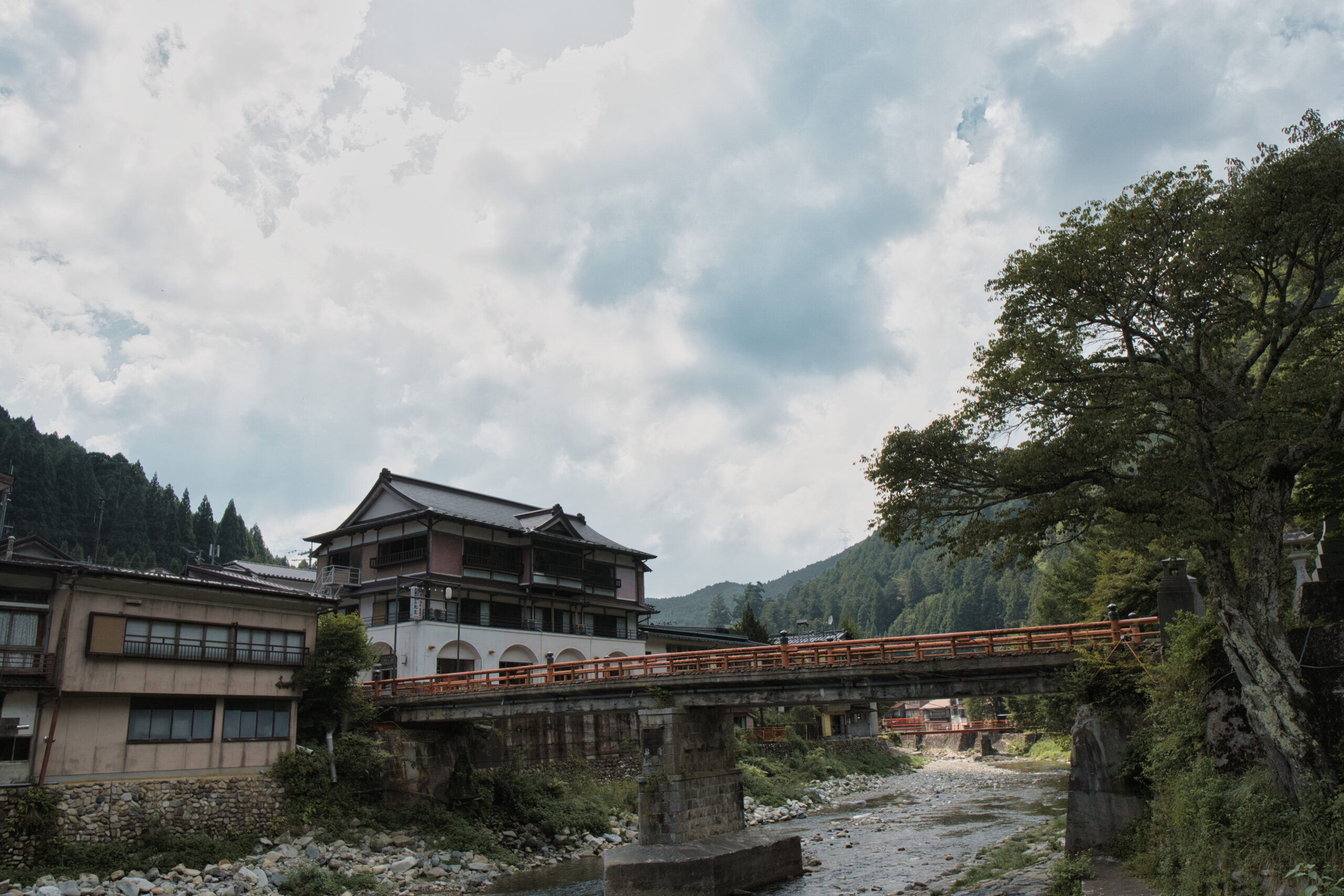 Traditional riverside scene with a multi-story building, red railing bridge, and rocky riverbank under a cloudy sky.