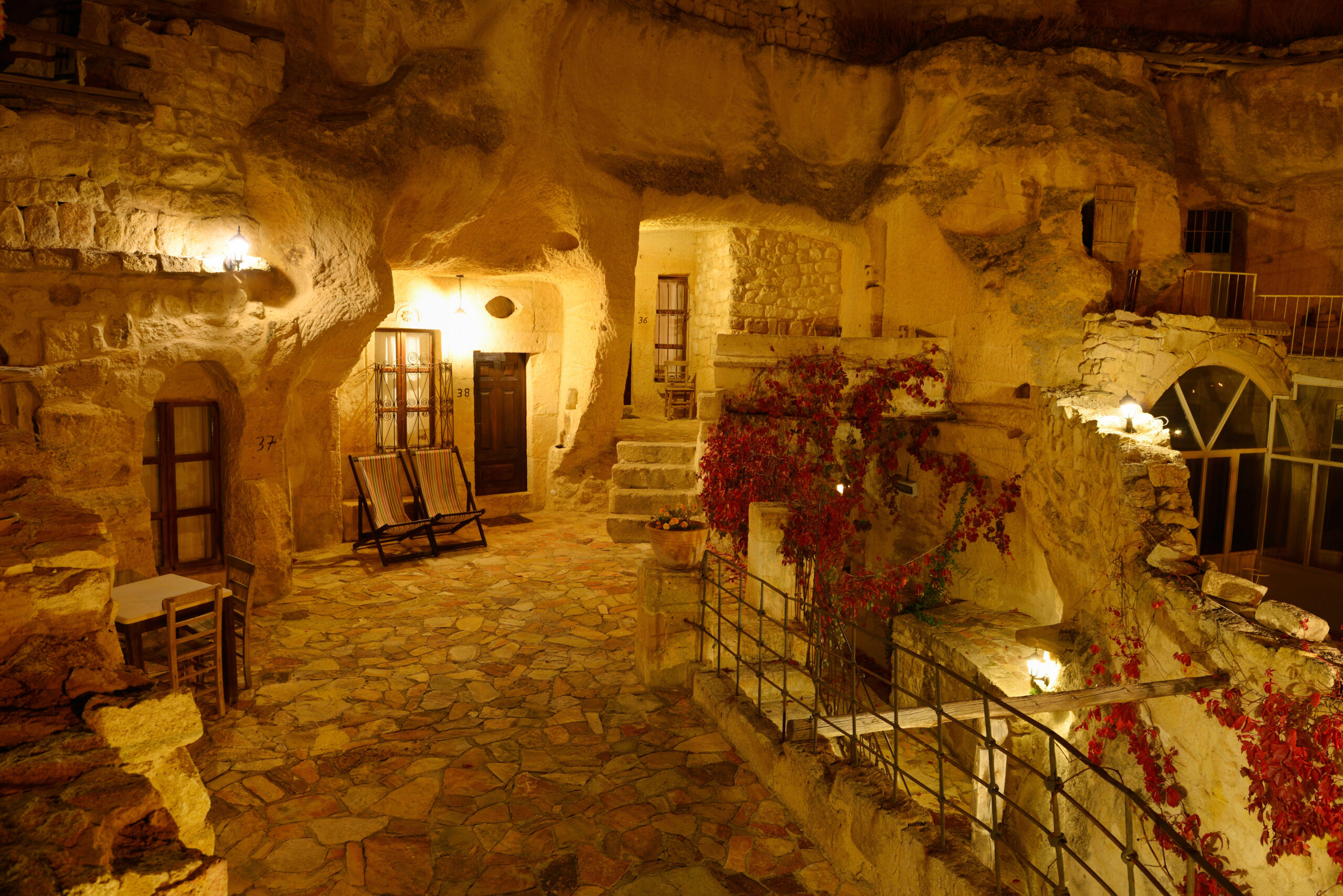 Warmly lit stone cavern courtyard at night with doors, windows, and outdoor seating amid ivy and rugged walls