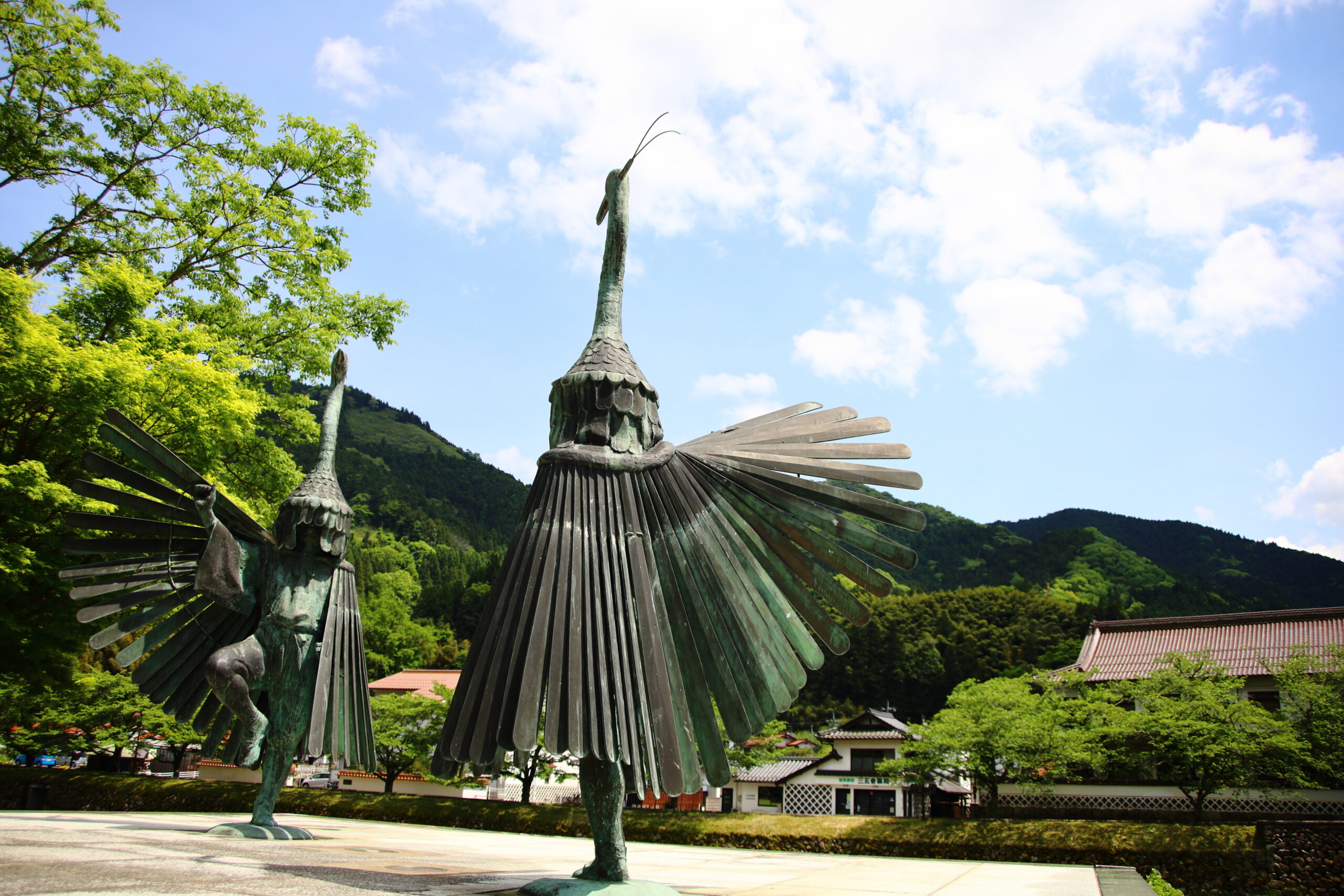 Bronze bird statue with outstretched wings in a sunny park, green trees and mountains in the background