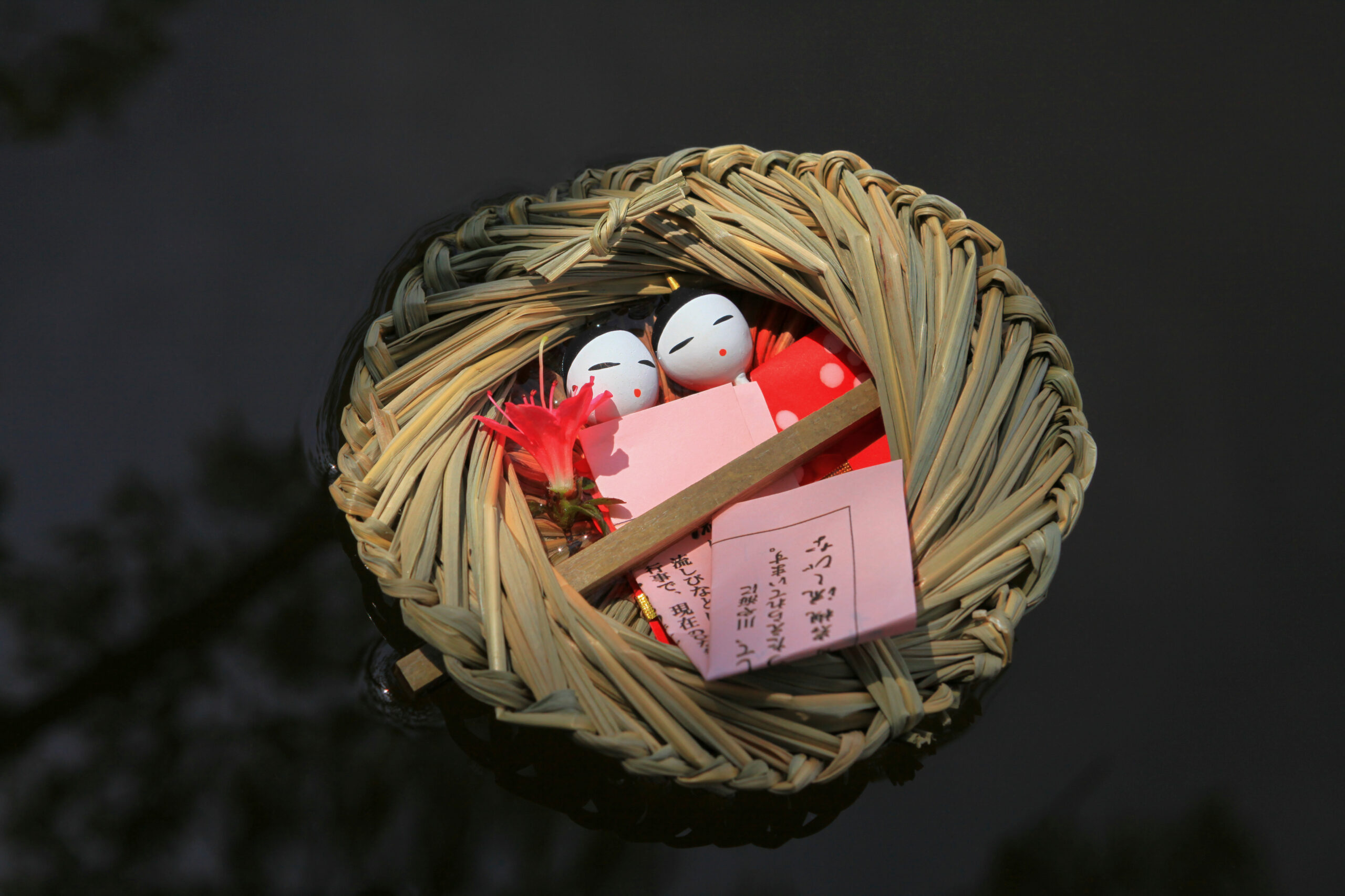Wicker basket wreath containing small white-faced Japanese dolls, pink cards, and red fabric on a dark surface.
