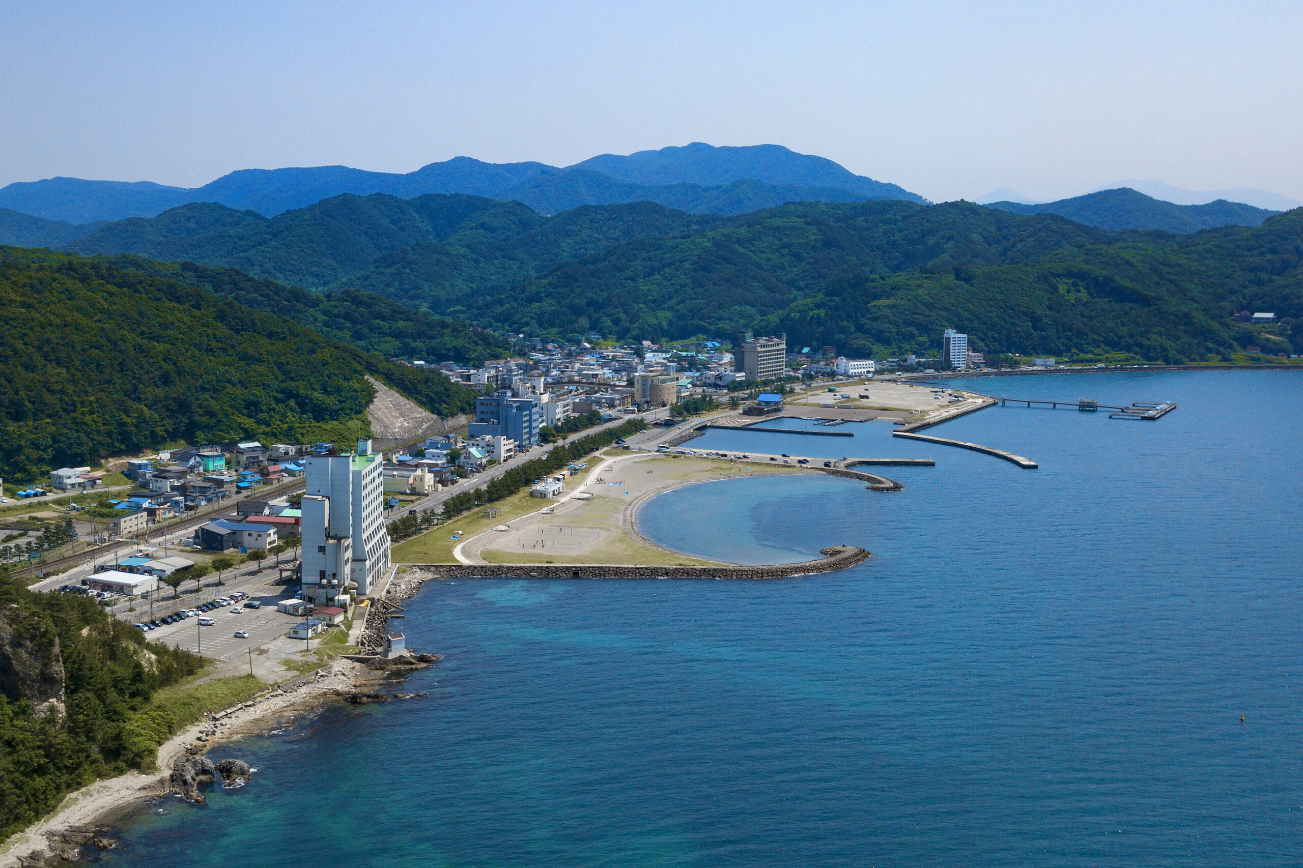 Coastal town along a curved beach with piers, low-rise buildings, and forested hills in the background.