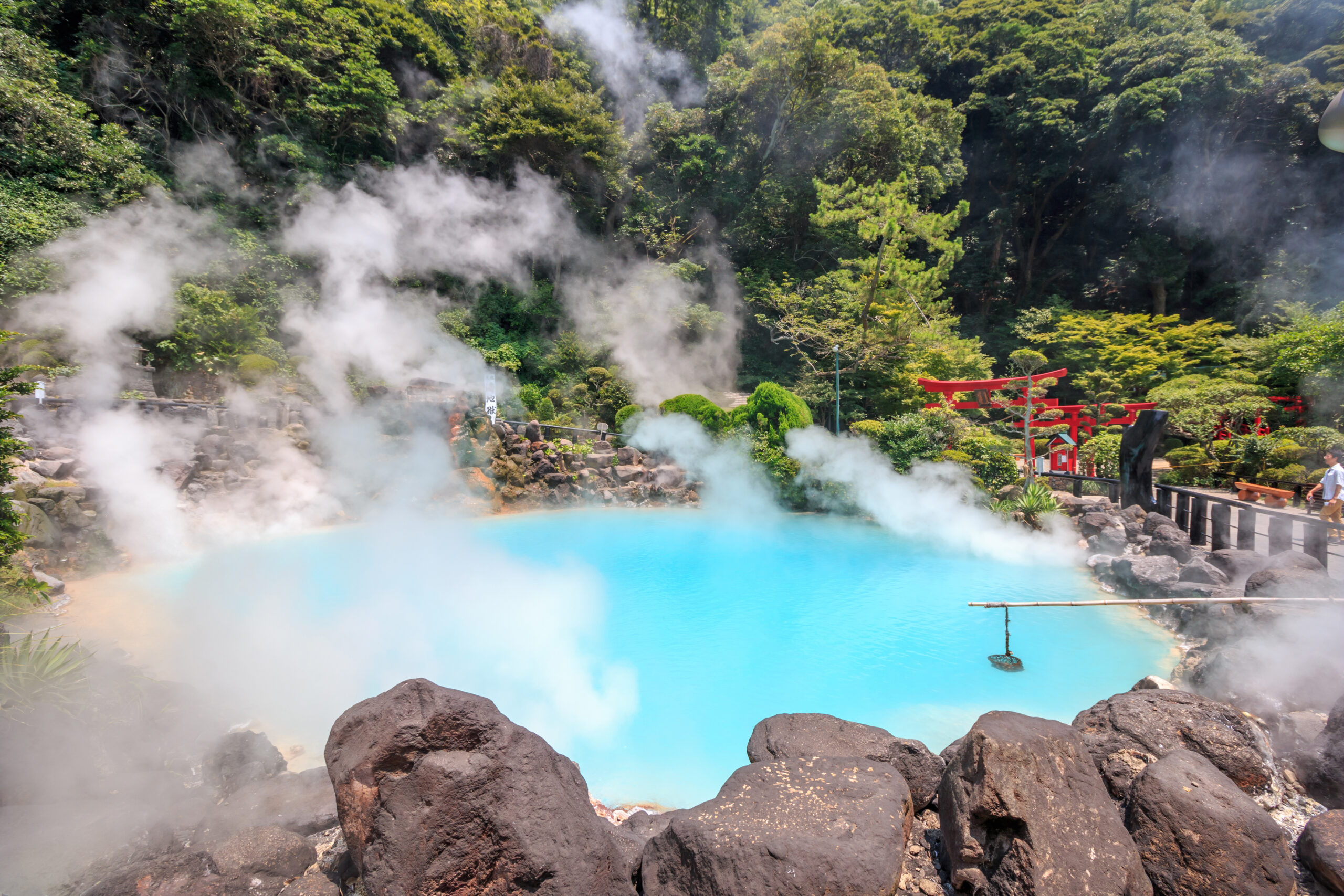 Steam rising from a bright blue hot spring, surrounded by rocks and dense forest, with red torii gates visible in the background.