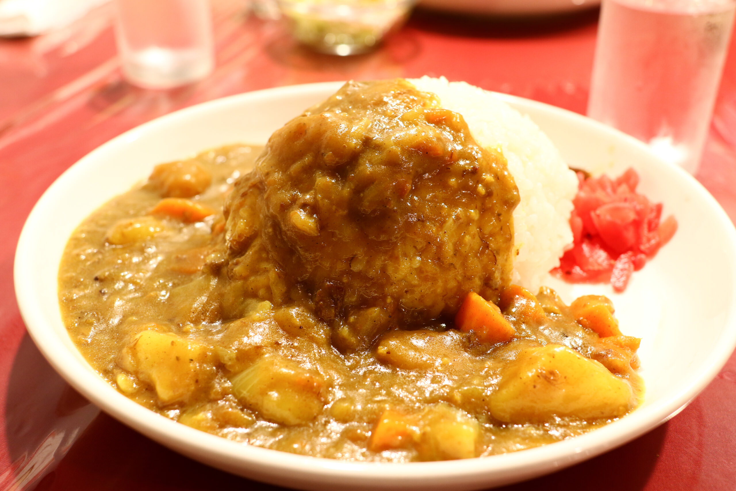 Bowl of creamy curry with rice and chopped vegetables, served on a red table with pickled ginger on the side.