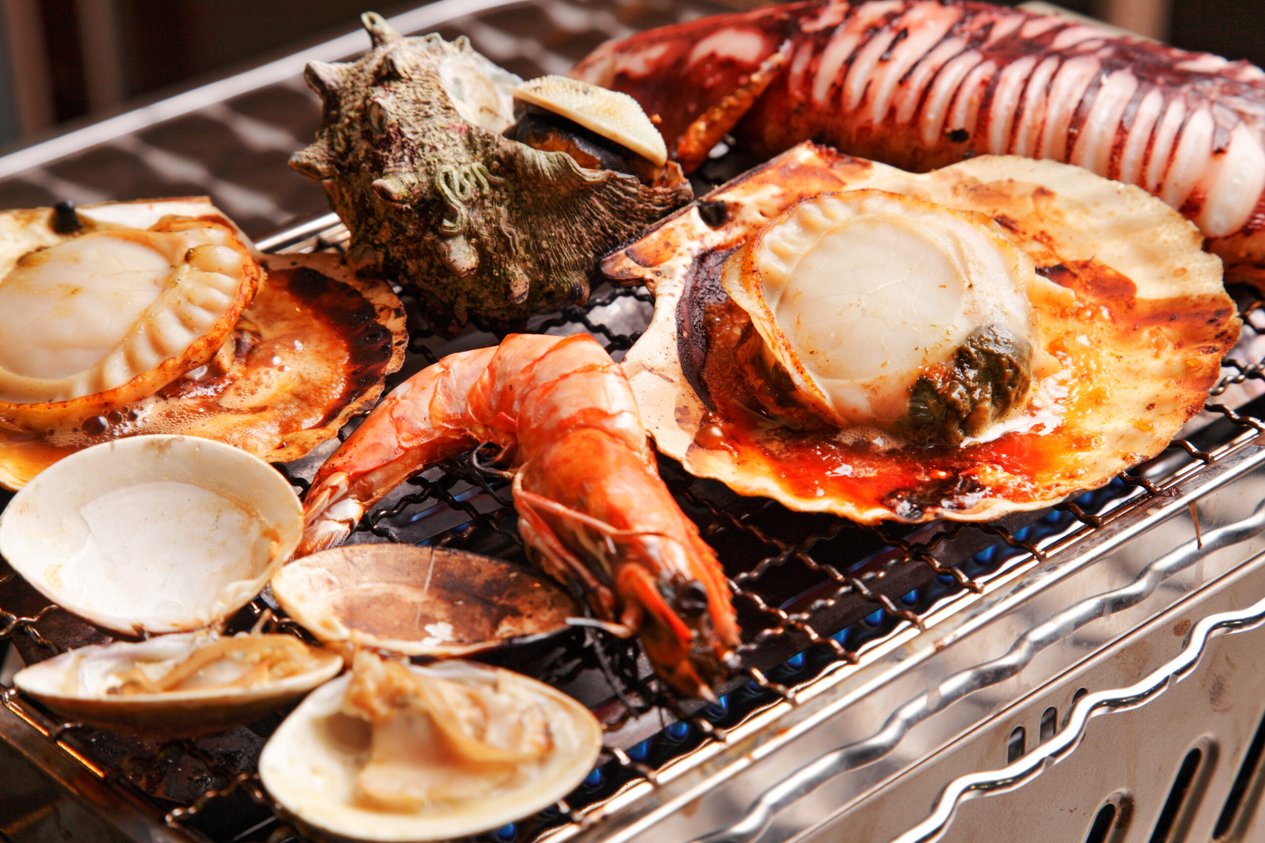 Assorted grilled seafood on a barbecue grill, including scallops in shells and shellfish with charred shells and sauce.