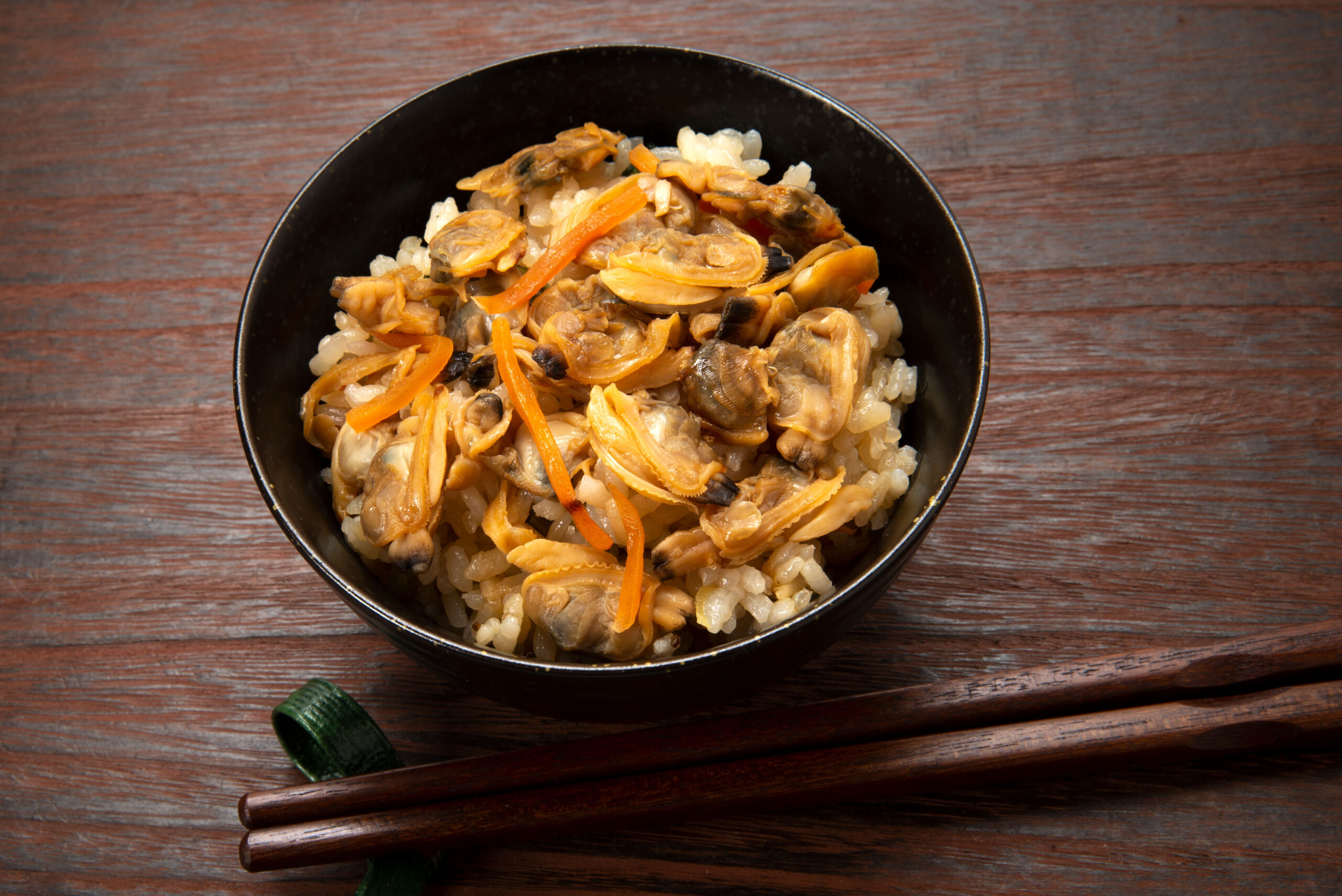 Bowl of white rice topped with mussels and orange carrot strips on a dark bowl, set on a wooden table with chopsticks nearby