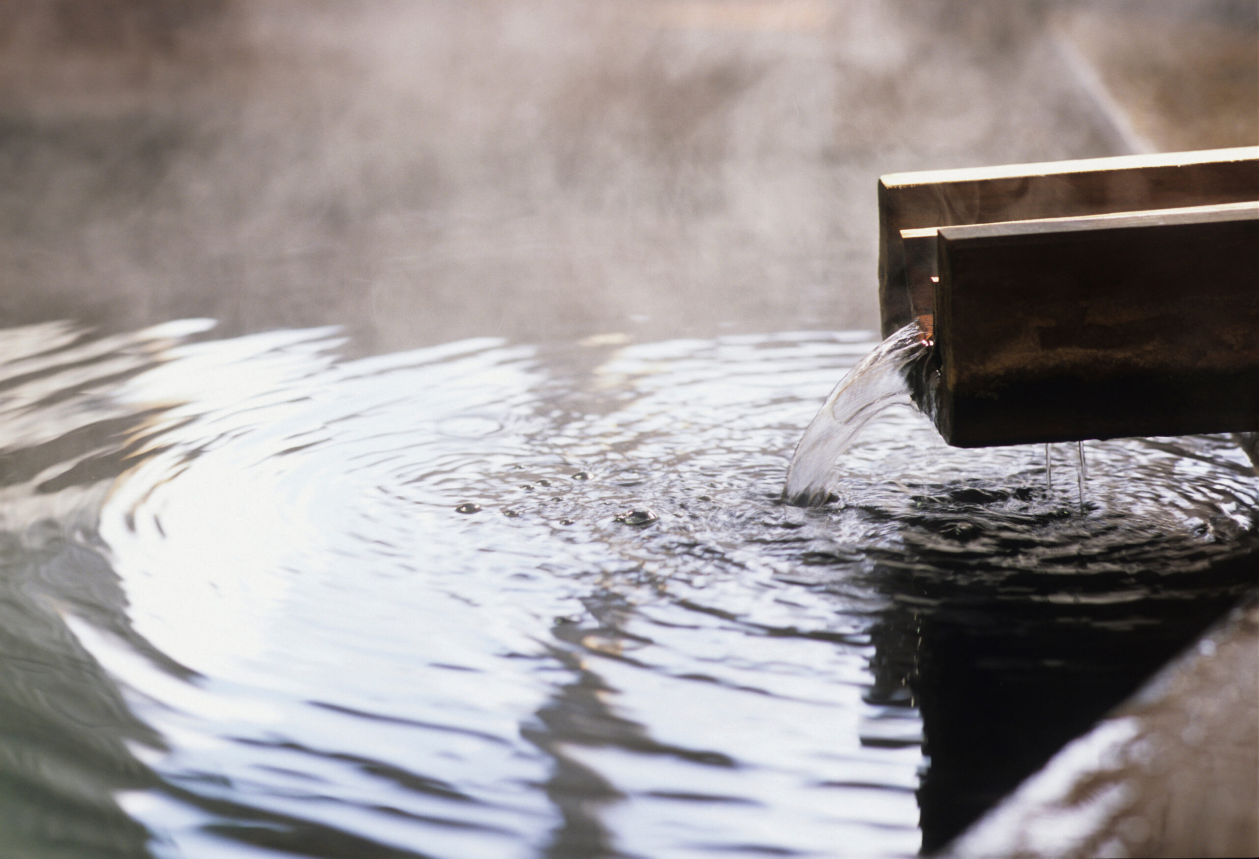 Wooden water trough pouring water into a rippling, misty pool with steam rising in the background and warm light.