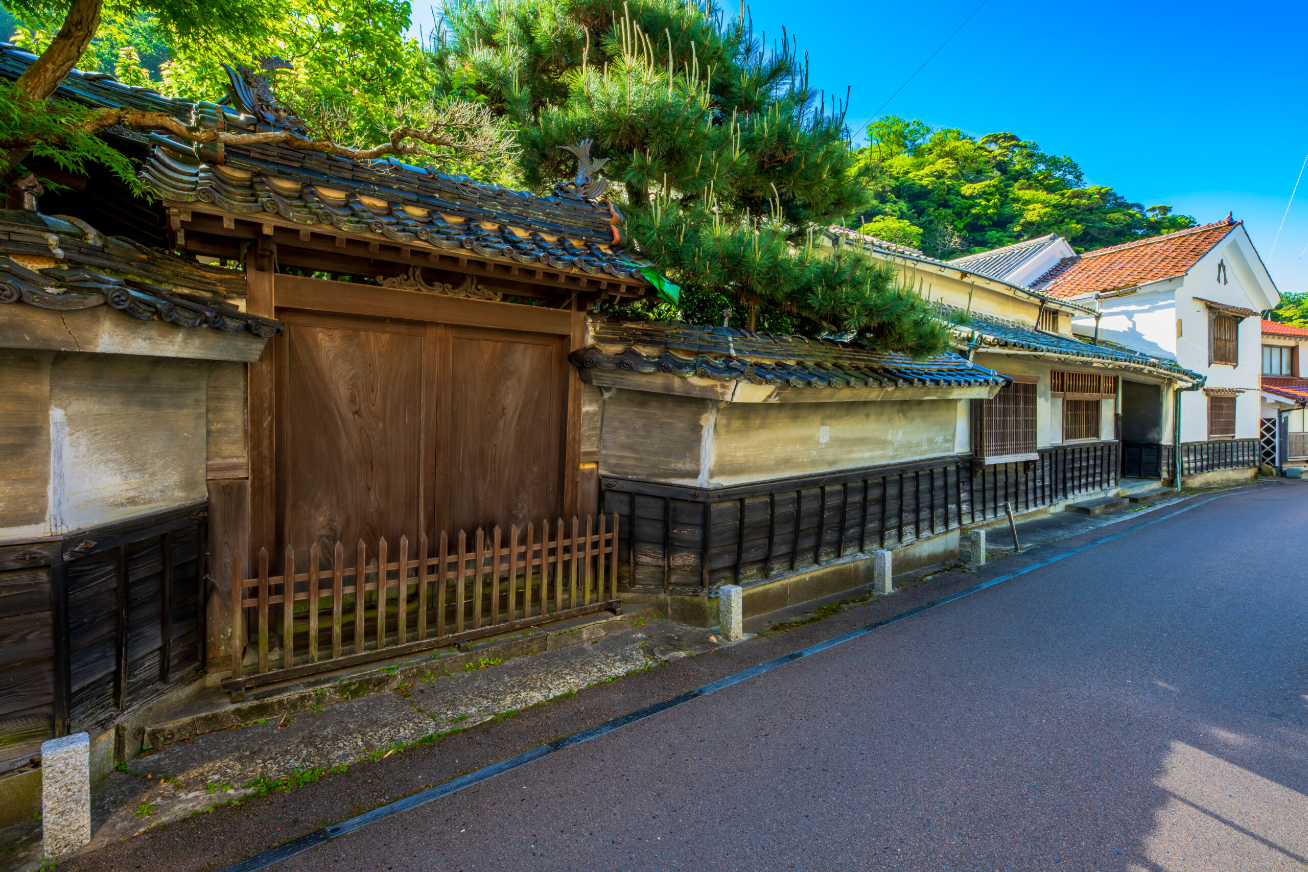 Traditional wooden gate and tile-walled fence along a quiet street with green trees above.,
