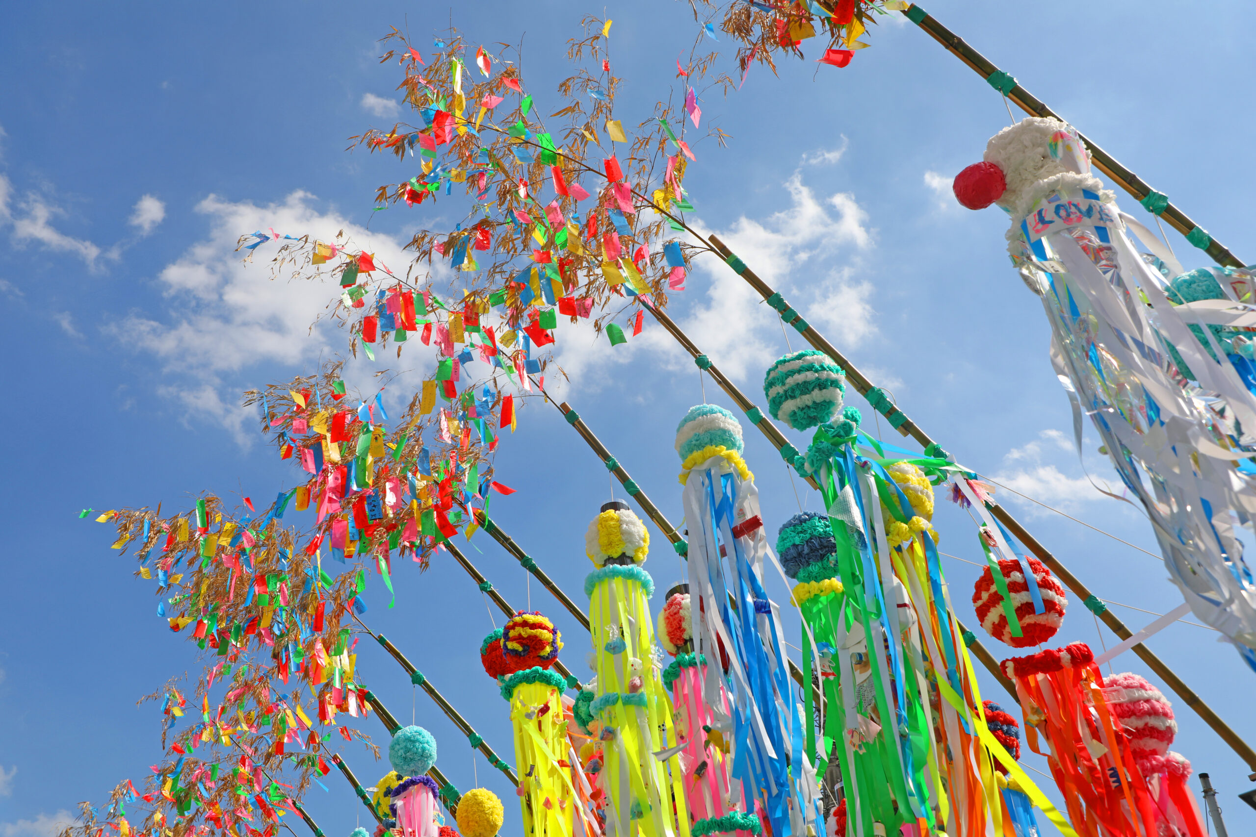 Bright festival decorations with multicolored streamers and puffy ornaments fluttering on bamboo poles against a blue sky.