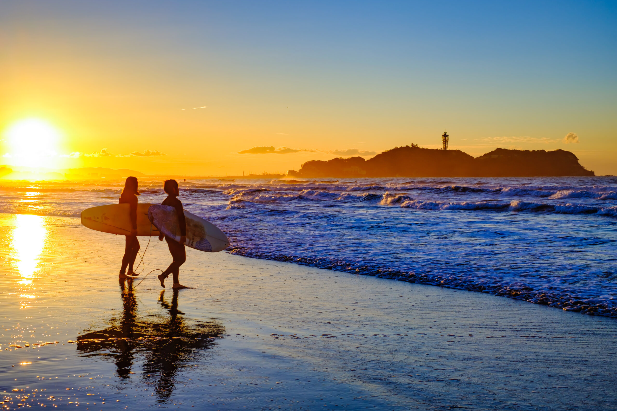 Two surfers carry their boards along the shoreline at sunset, with a distant island and lighthouse visible on the horizon.