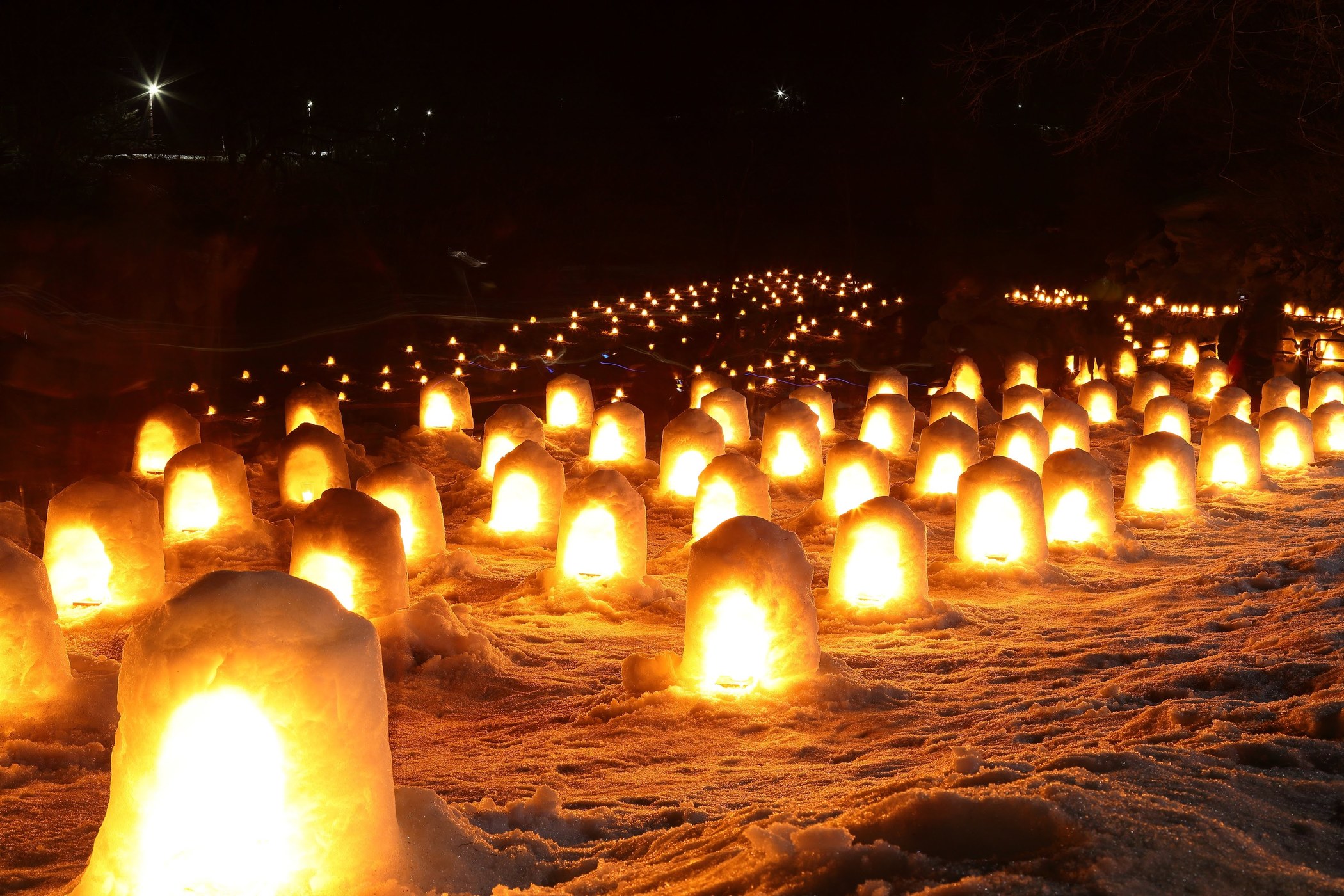 Row of glowing lanterns arranged in the snow at night, casting a warm amber light across a dark landscape.