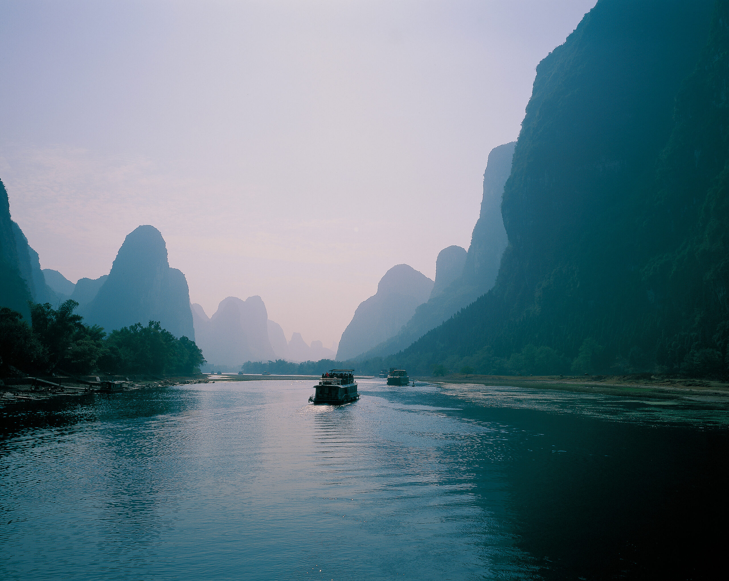 Boat cruising along a calm river flanked by towering misty karst mountains on both sides.