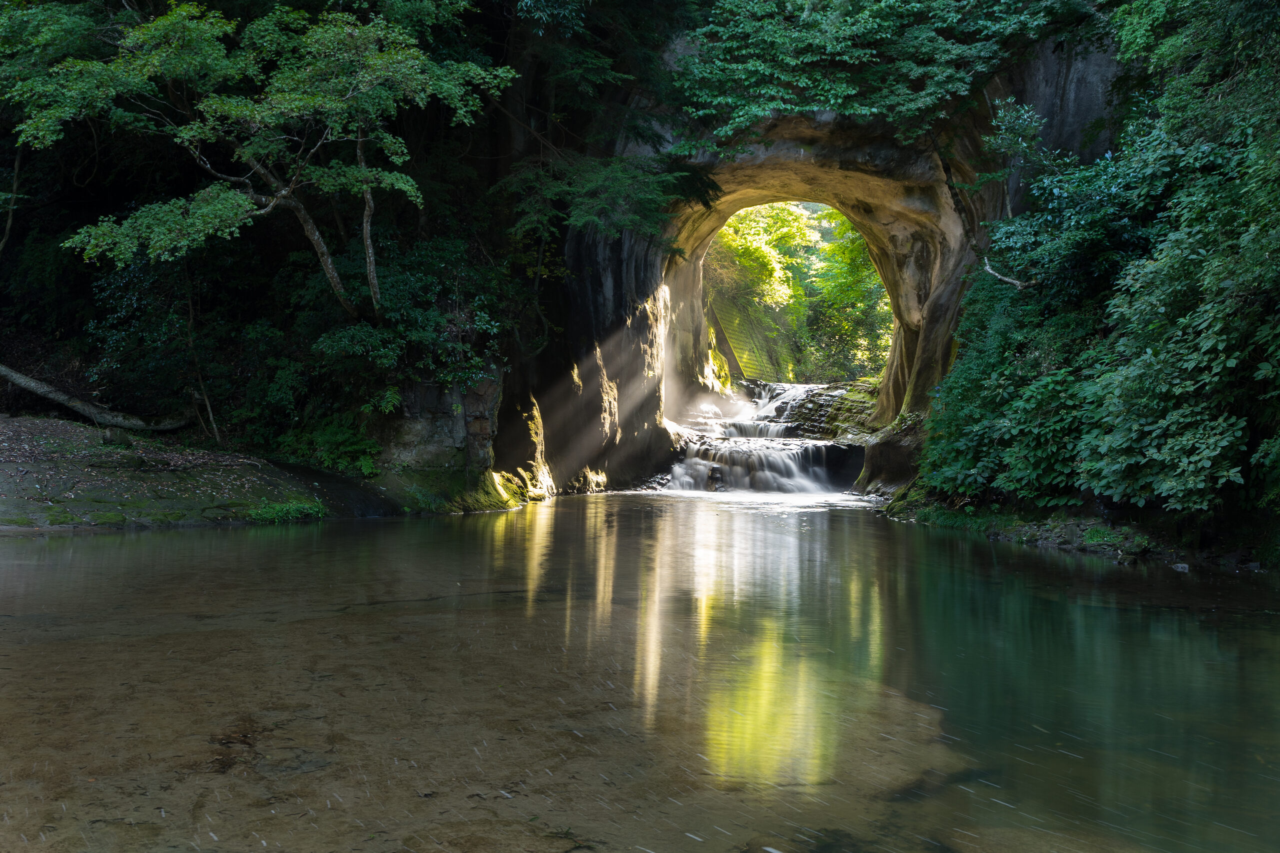 Sunlit natural arch over a small waterfall in a forest, reflecting in calm water on a tranquil pool.