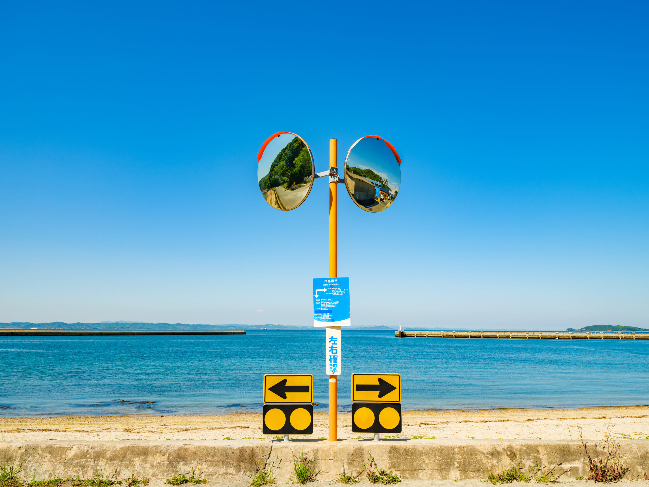 Orange traffic post with two circular convex mirrors reflecting a coastal road and green trees against a clear blue sky.