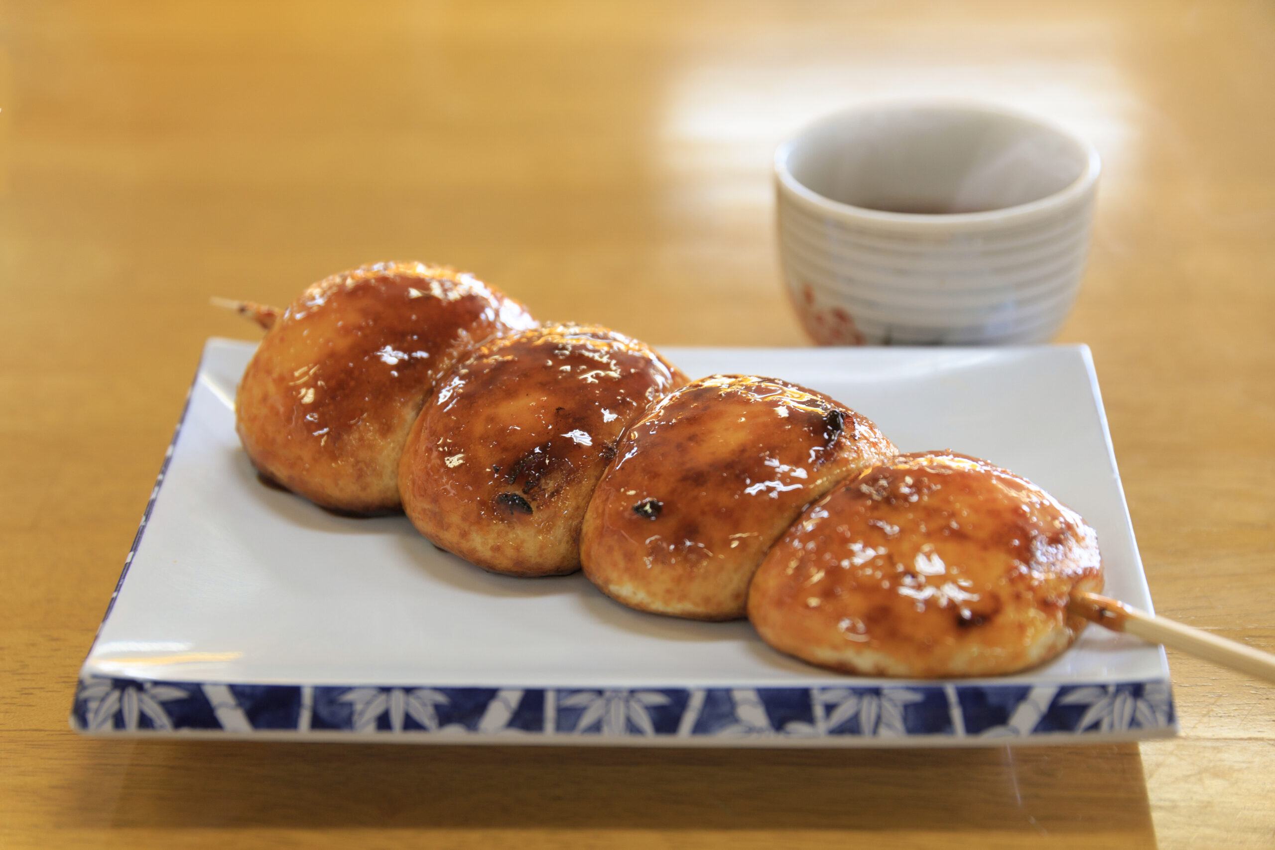 Glazed buns on a white plate with a blue patterned edge, placed on a wooden table, with a white ceramic cup in the background.