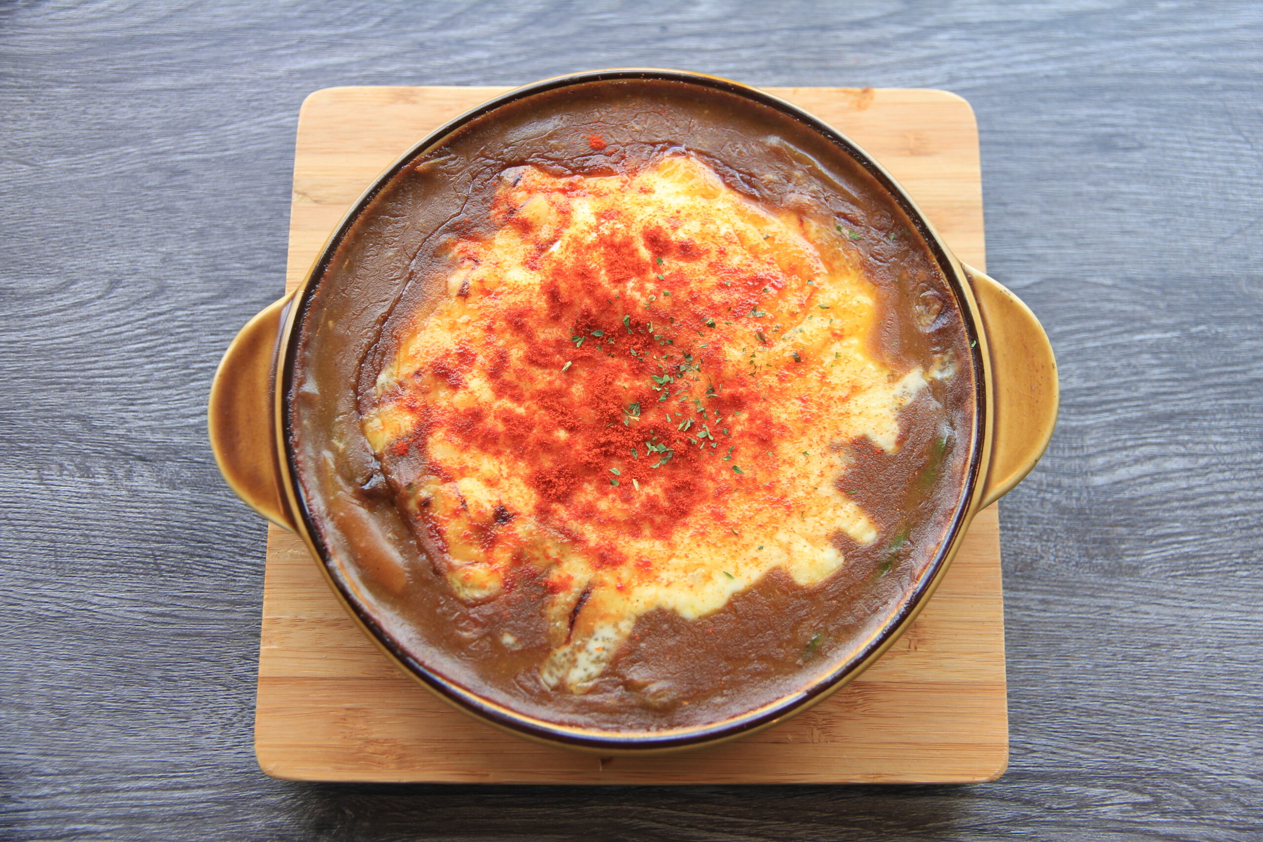 Oven-baked cheese-topped casserole in a round brown dish on a wooden board, with red seasoning sprinkled on top.