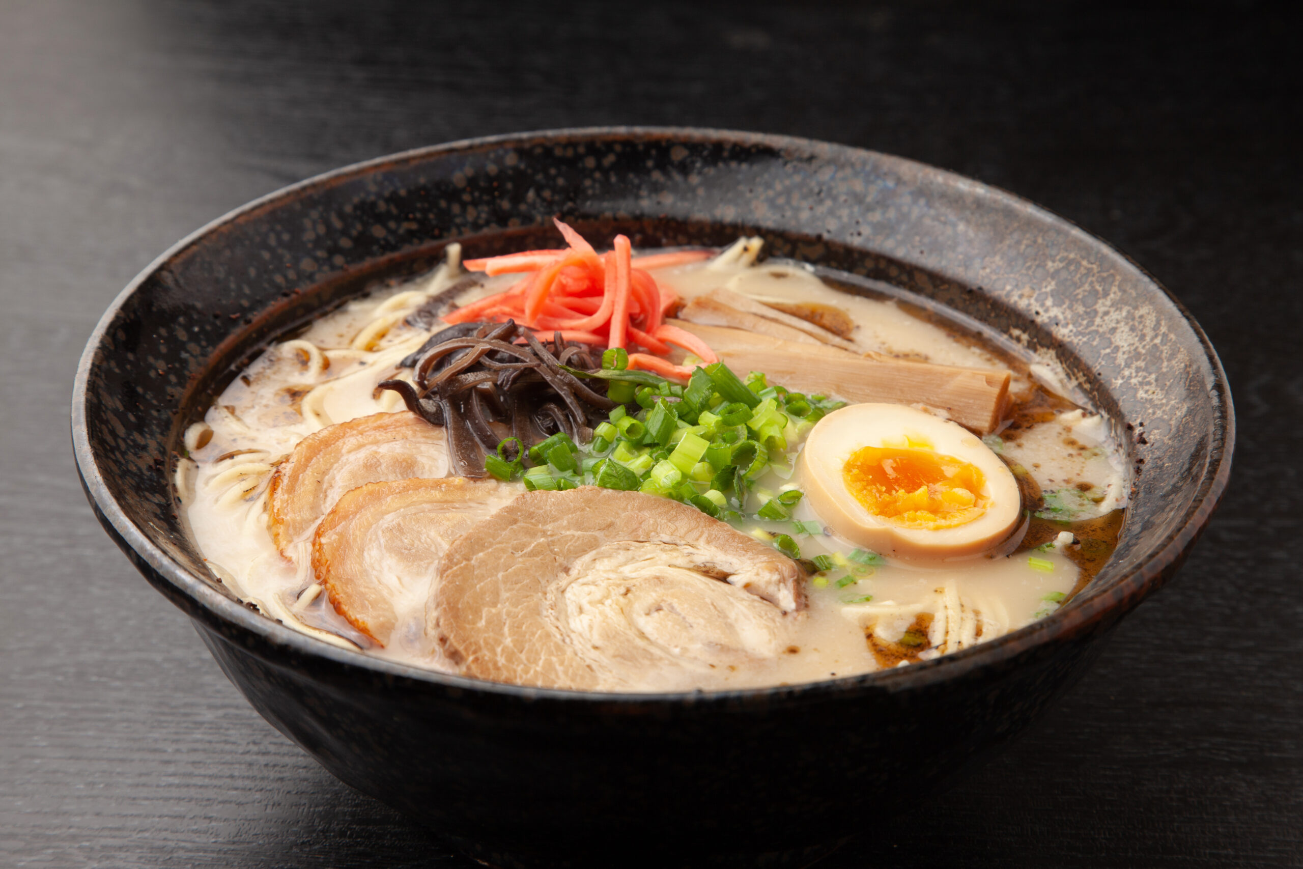 Bowl of ramen with pork slices, half of a soft-boiled egg, green onions, pickled ginger, and noodles in a creamy broth.