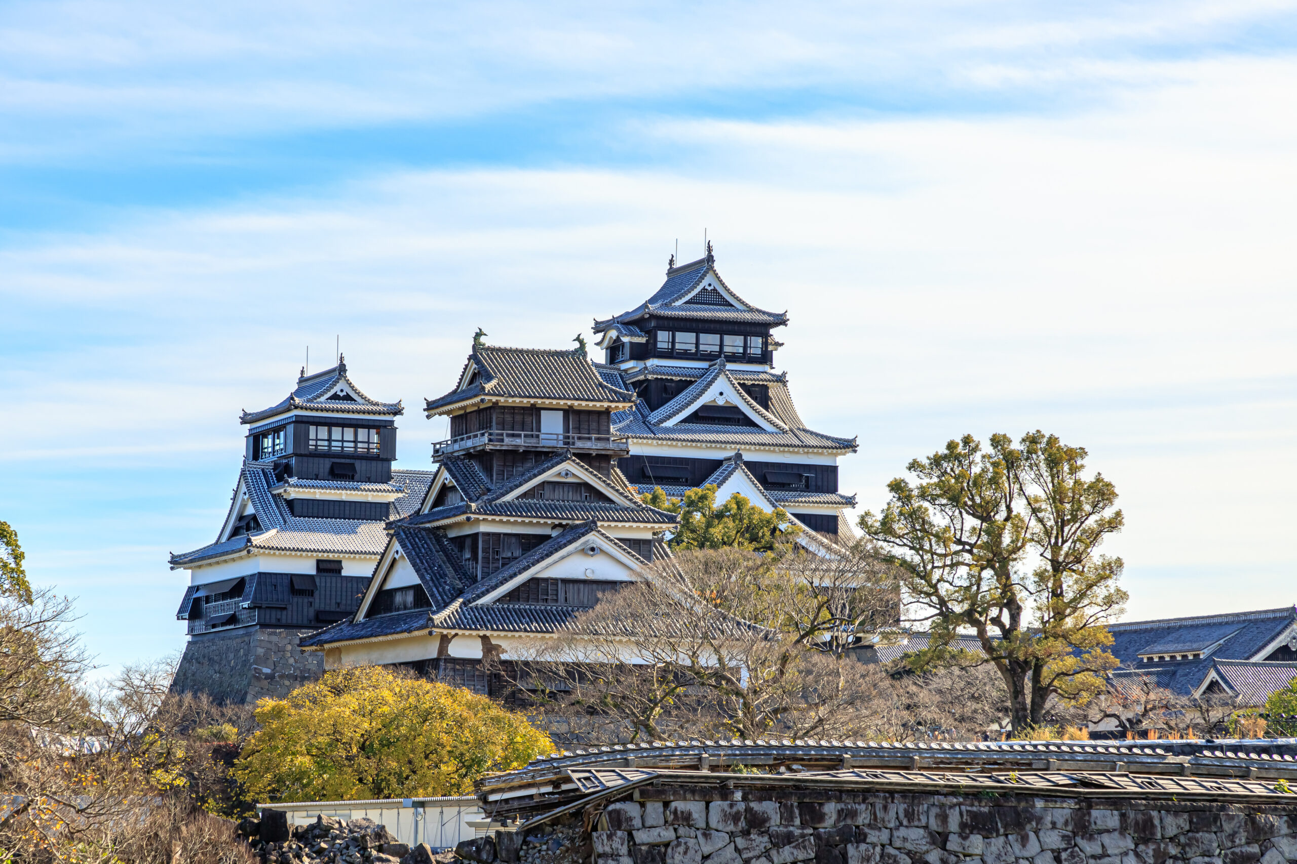 Traditional Japanese castle with blue-tiled roofs on a stone hill, surrounded by trees under a clear blue sky.