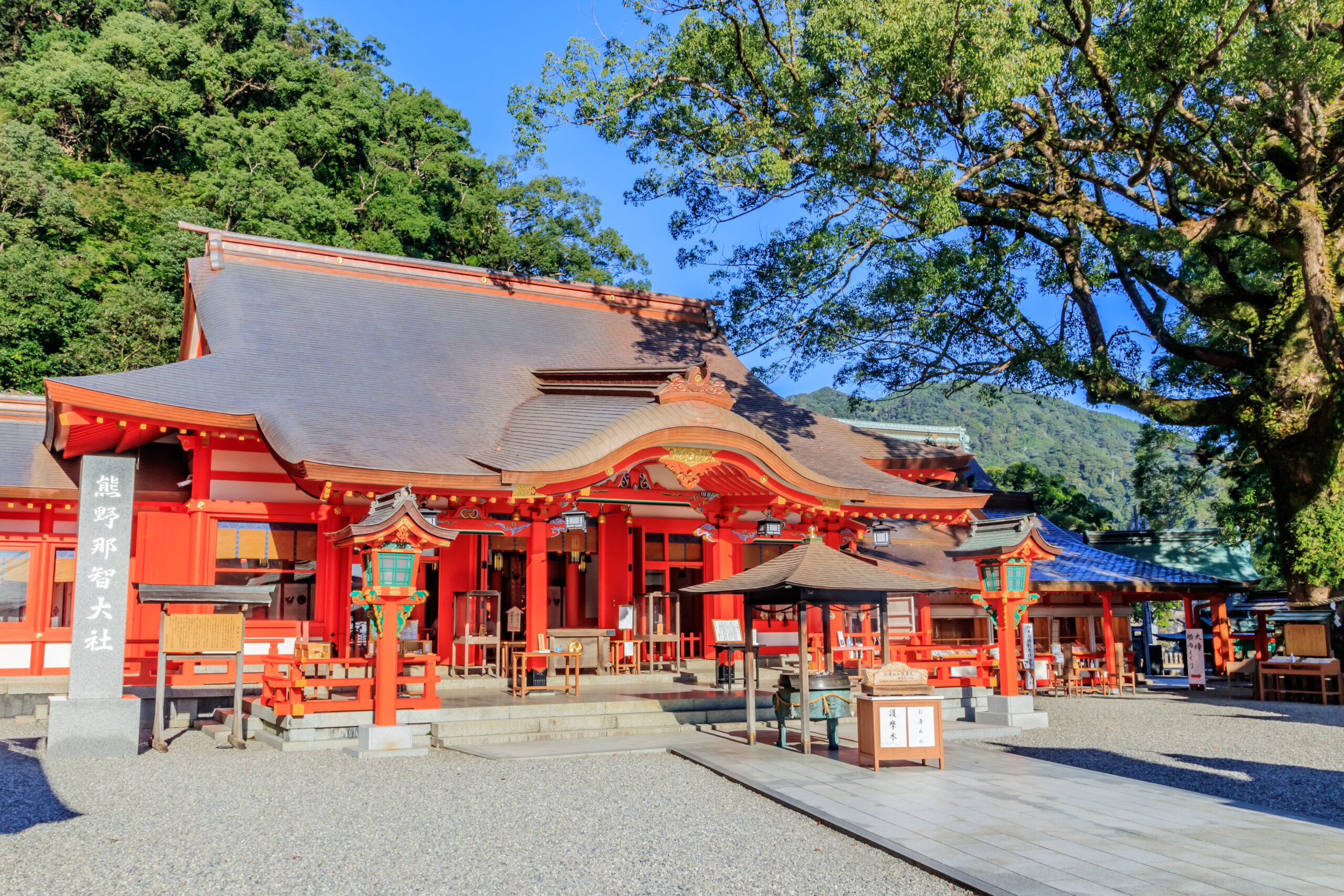 Shinto shrine with bright red buildings, ornate wooden carvings, and a gravel courtyard surrounded by green trees under a clear blue sky