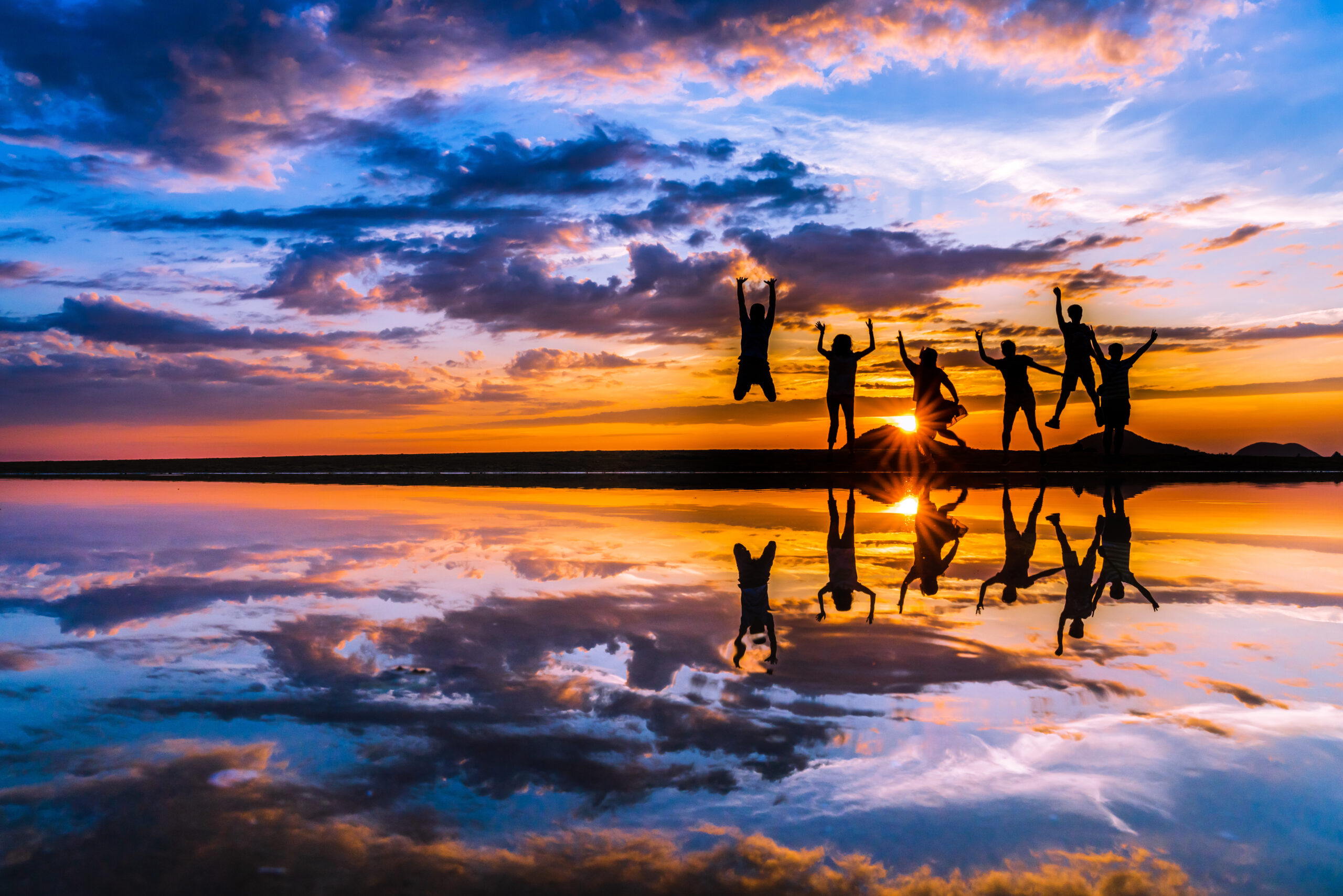 Silhouetted group of friends jumping with arms raised at sunset on a beach, reflected in calm water with a colorful sky.